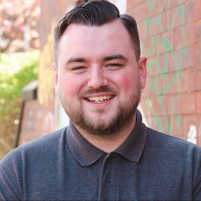 A smiling young man with short dark hair and a beard, standing outdoors next to a graffiti-covered brick wall.