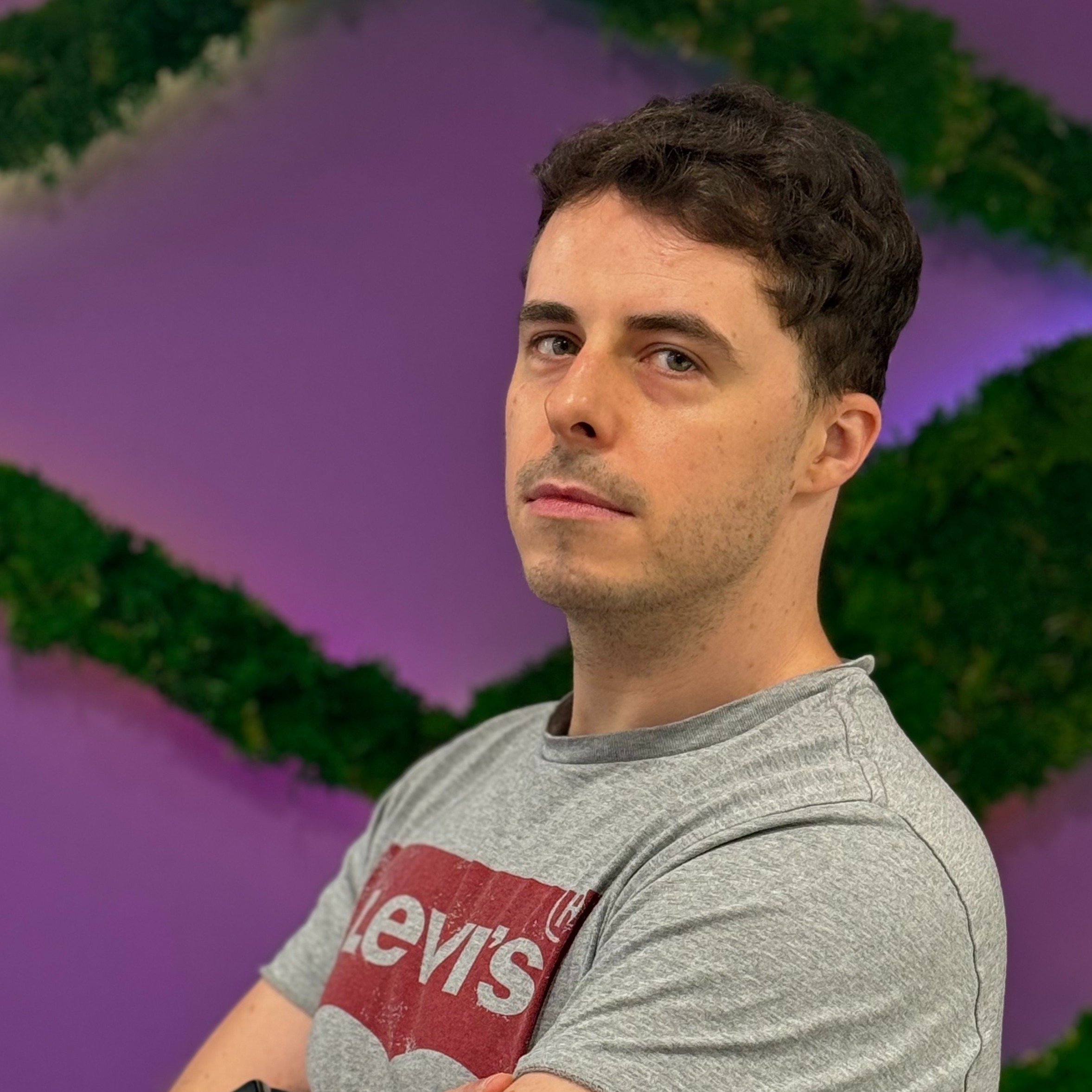 A young man with short dark hair and light skin, wearing a gray Levi's T-shirt, stands with arms crossed in front of a colorful abstract background with purple and green patterns.