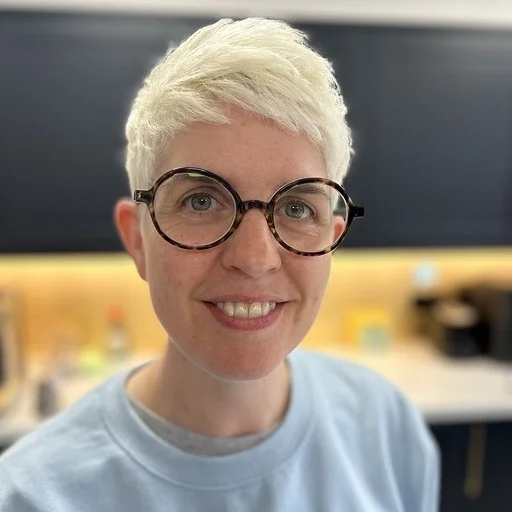 A woman with short platinum blonde hair, wearing large round glasses and a light blue shirt, smiling indoors.