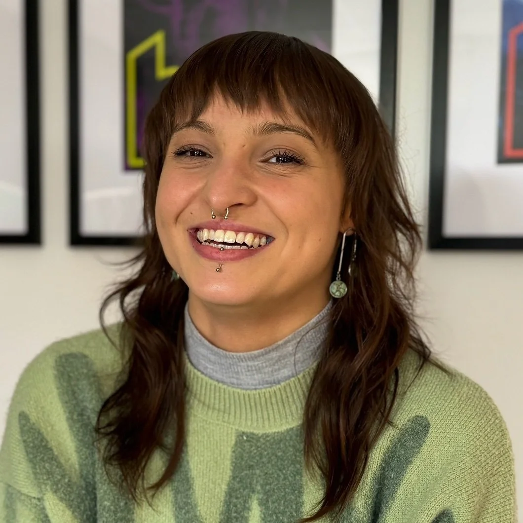 A young woman with brown hair, smiling with braces, wearing a green sweater and a gray turtleneck, in front of framed colorful artwork.