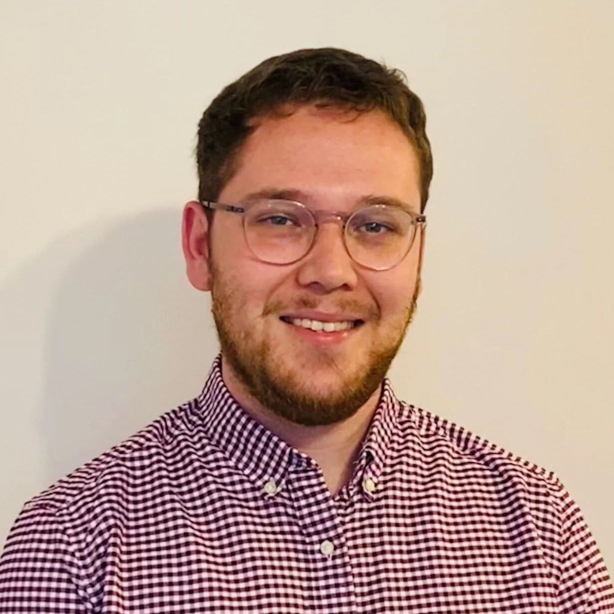 A young man with short brown hair, glasses, and a beard, smiling, wearing a red and white checkered shirt, standing against a plain white background.