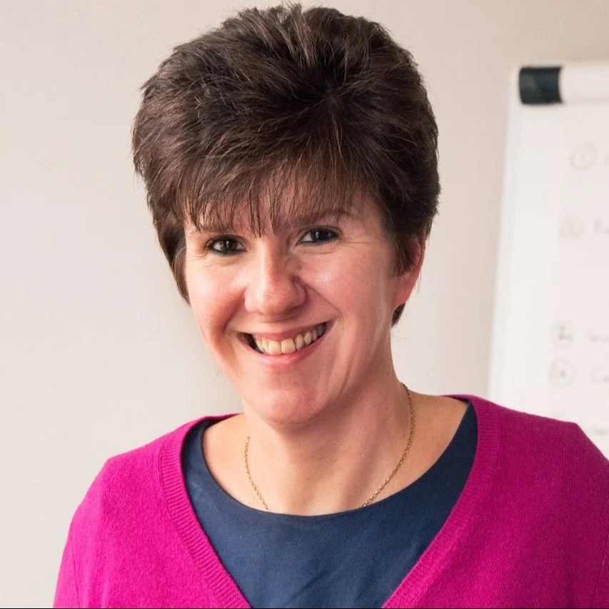 A woman smiling, wearing a pink sweater over a navy shirt, with short brown hair, standing in front of a whiteboard.