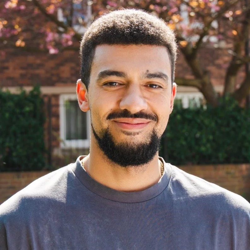 A young man with short curly hair and a beard, smiling outdoors on a sunny day, with pink flowering tree and brick building in the background.