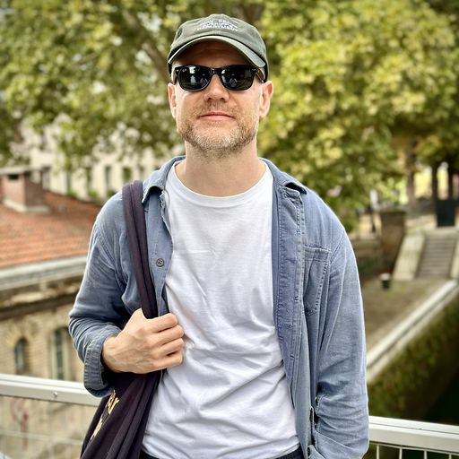 Man wearing sunglasses, baseball cap, denim shirt layered over a white t-shirt, and carrying a backpack, standing outdoors with trees and buildings in the background.
