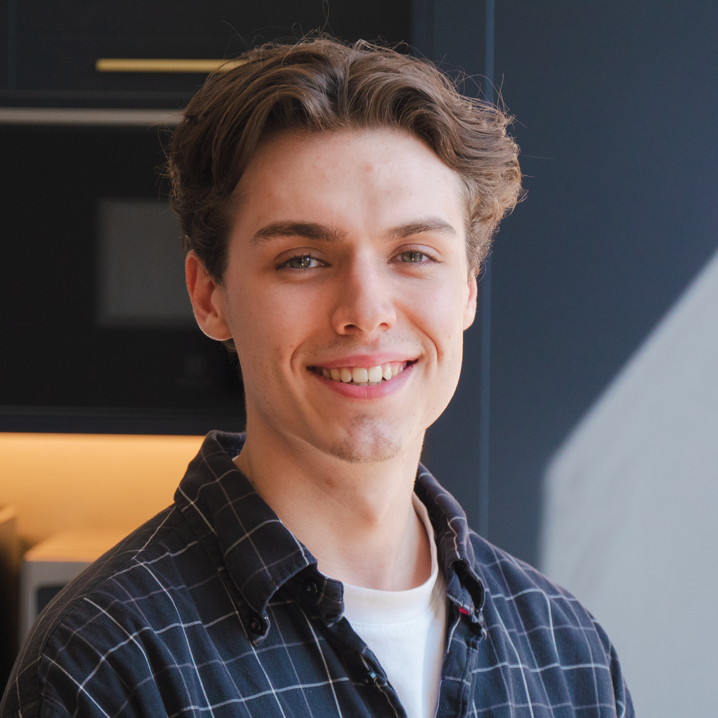 Close-up of a young man with light skin and brown hair smiling indoors.
