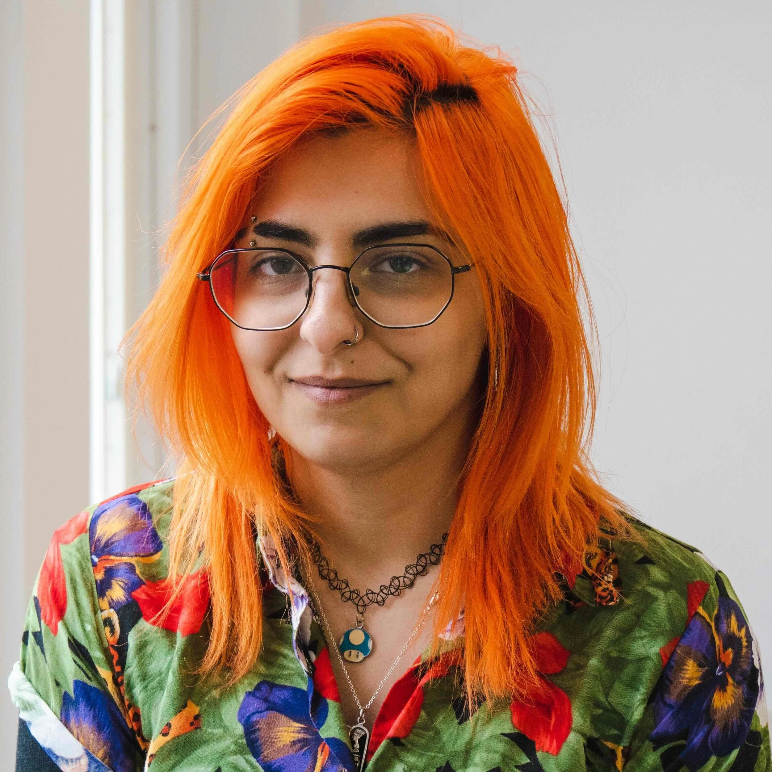 A woman with bright orange hair wearing glasses, a floral shirt, and layered necklaces, standing near a window with a neutral background.