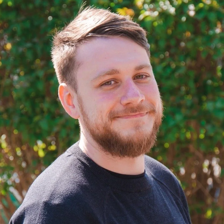 A young man with light skin, a beard, and short brown hair smiling outdoors with greenery in the background.
