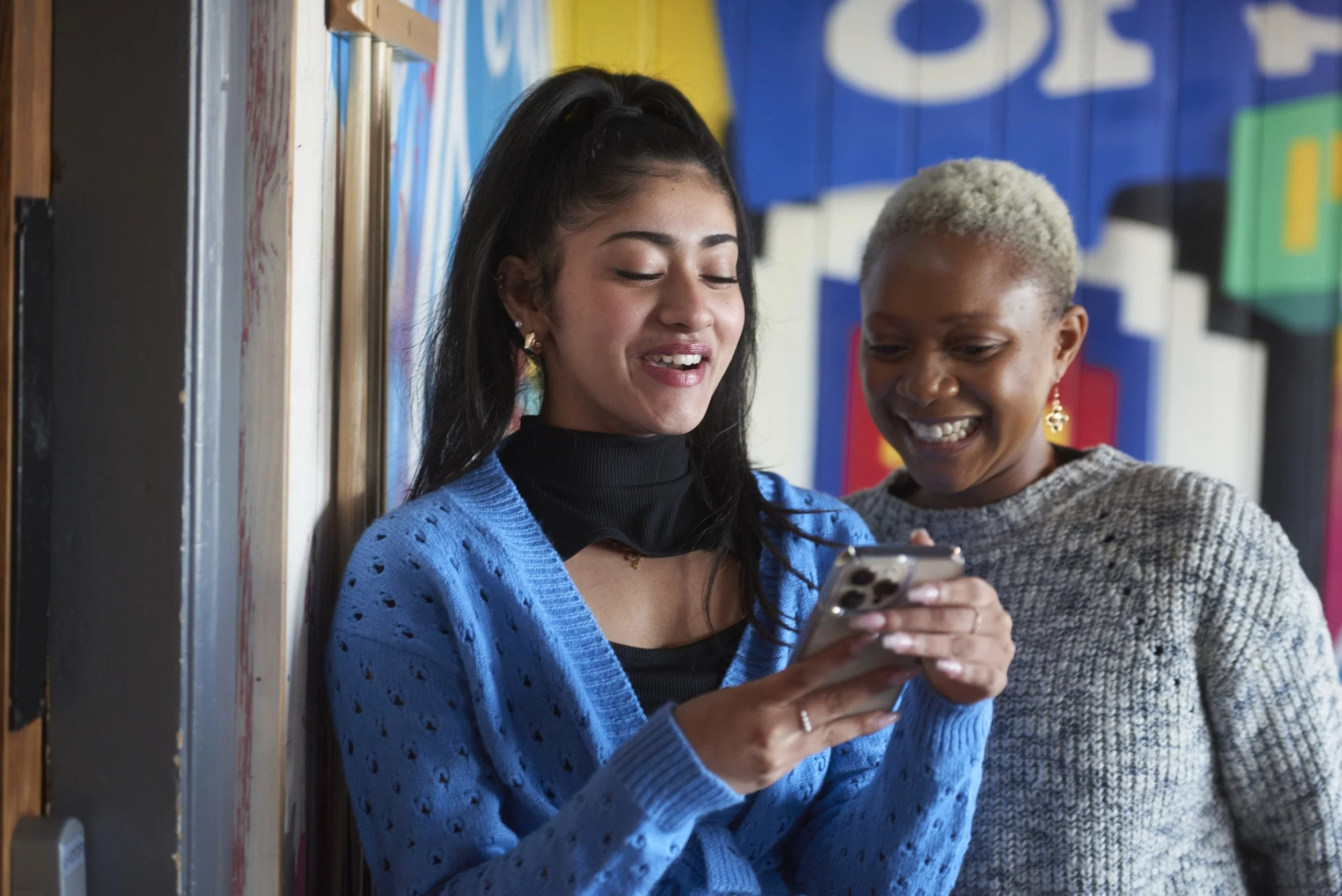 Two women smiling and looking at a smartphone together in an indoor setting with colorful wall art.