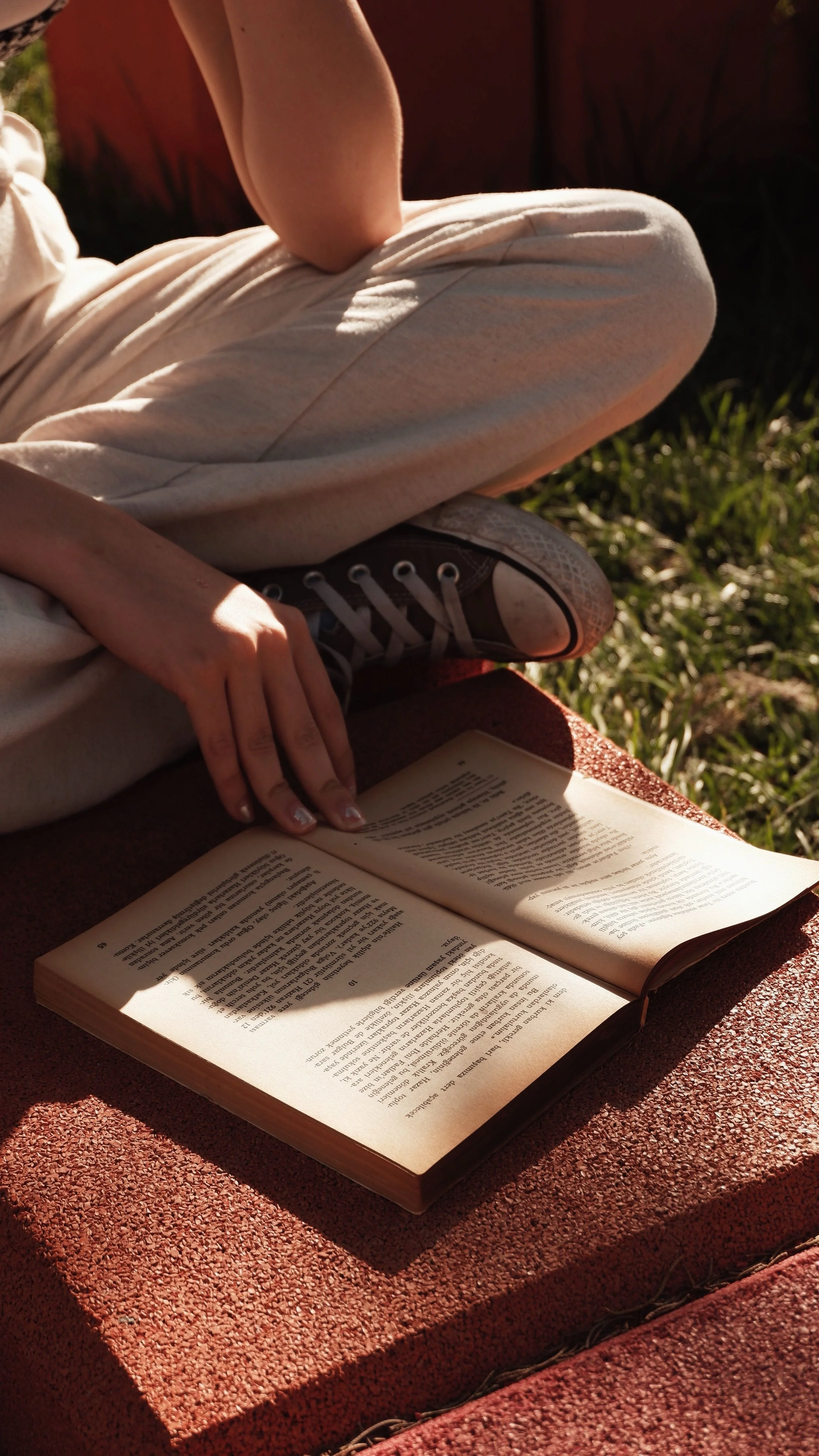 A person sitting on a red bench, reading a book outdoors in sunlight, wearing beige pants, a short sleeve shirt, and sneakers, with grass in the background.