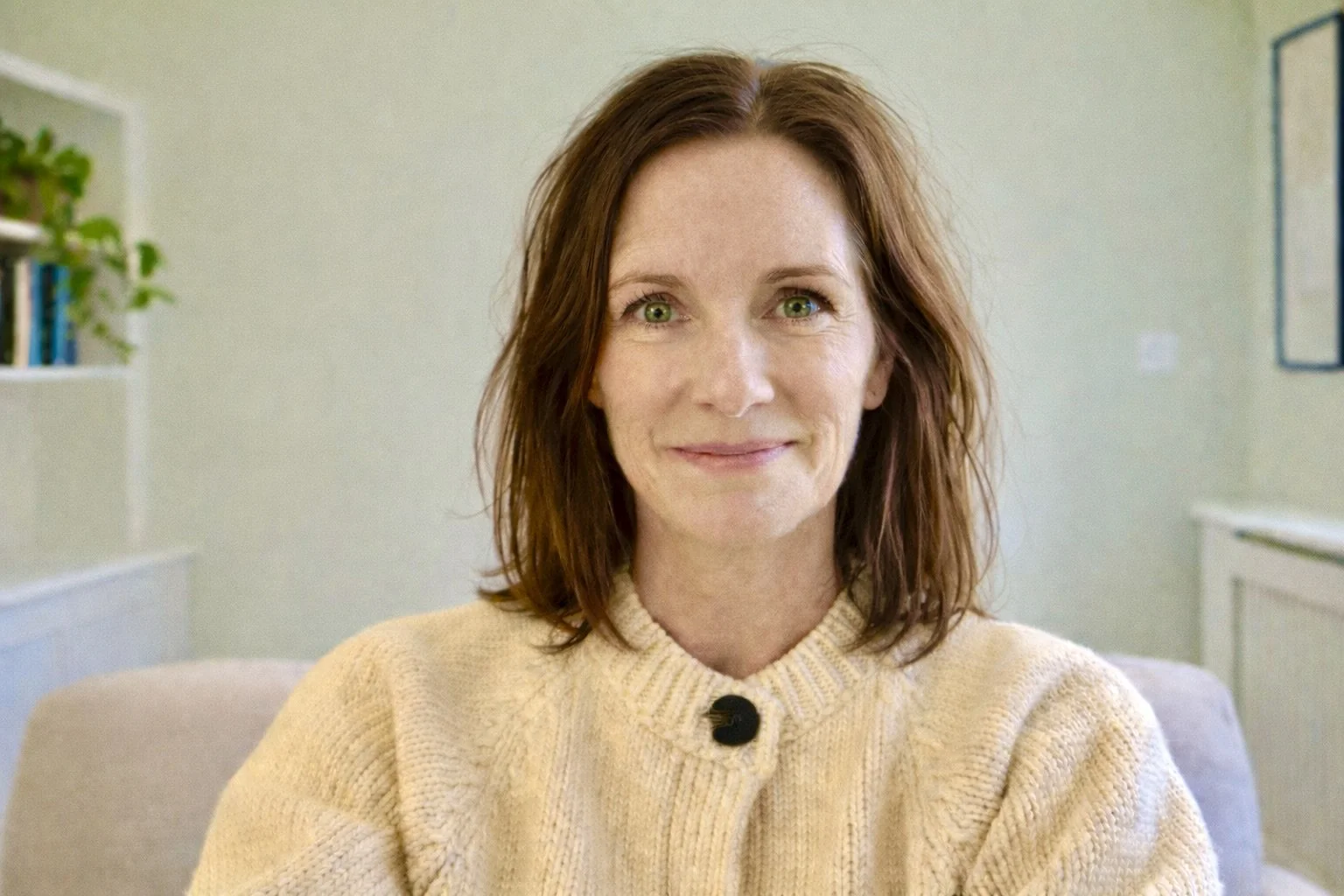 A woman with shoulder-length reddish-brown hair and green eyes smiling while sitting indoors wearing a cream-colored knit sweater.