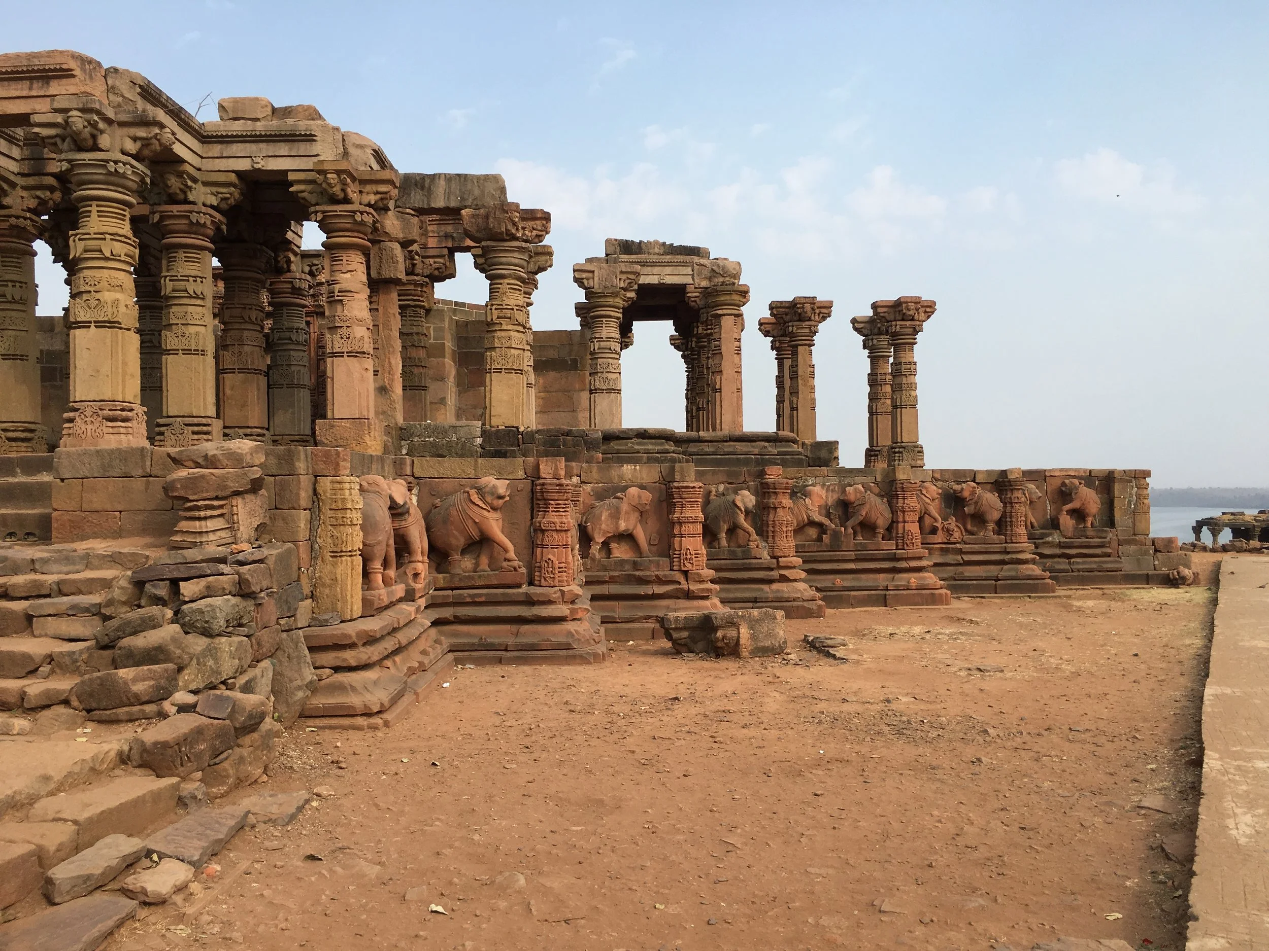 Ancient stone ruins with pillars, carvings, and statues of elephants, set on a dirt ground near a body of water.