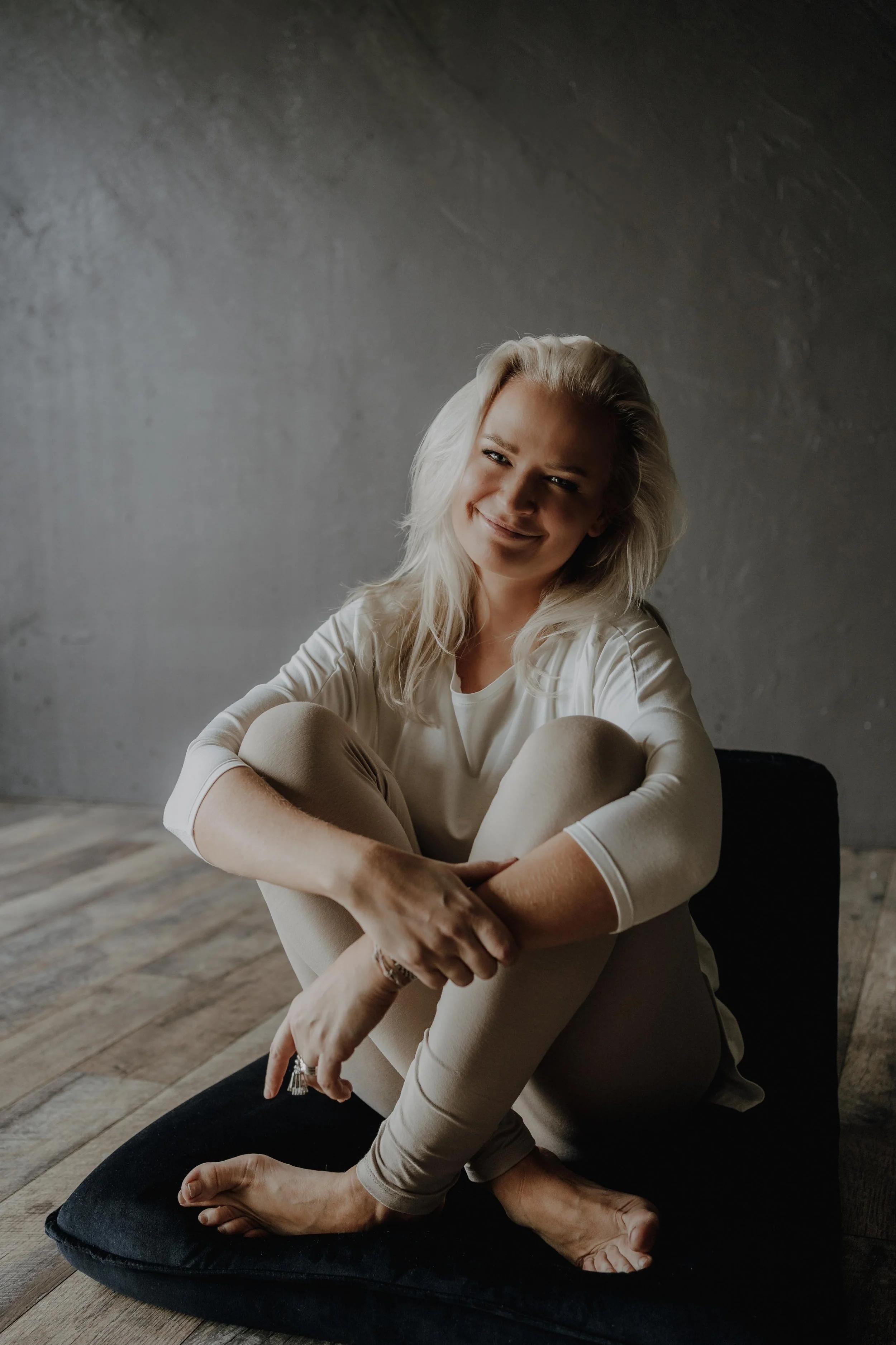 A woman with blonde hair sitting on a black cushion on a wooden floor, smiling, with a gray wall behind her.
