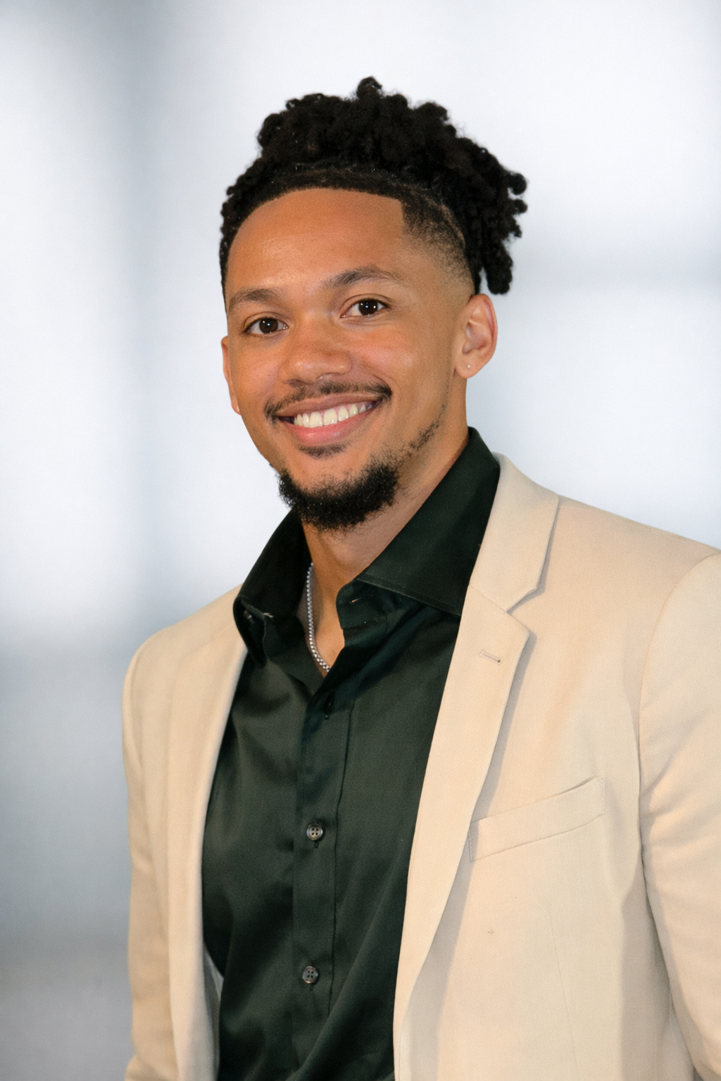 A smiling man with curly hair styled in a bun, a goatee, and a small earring in his left ear, wearing a beige blazer over a black shirt with a collar, posing against a plain light gray background.