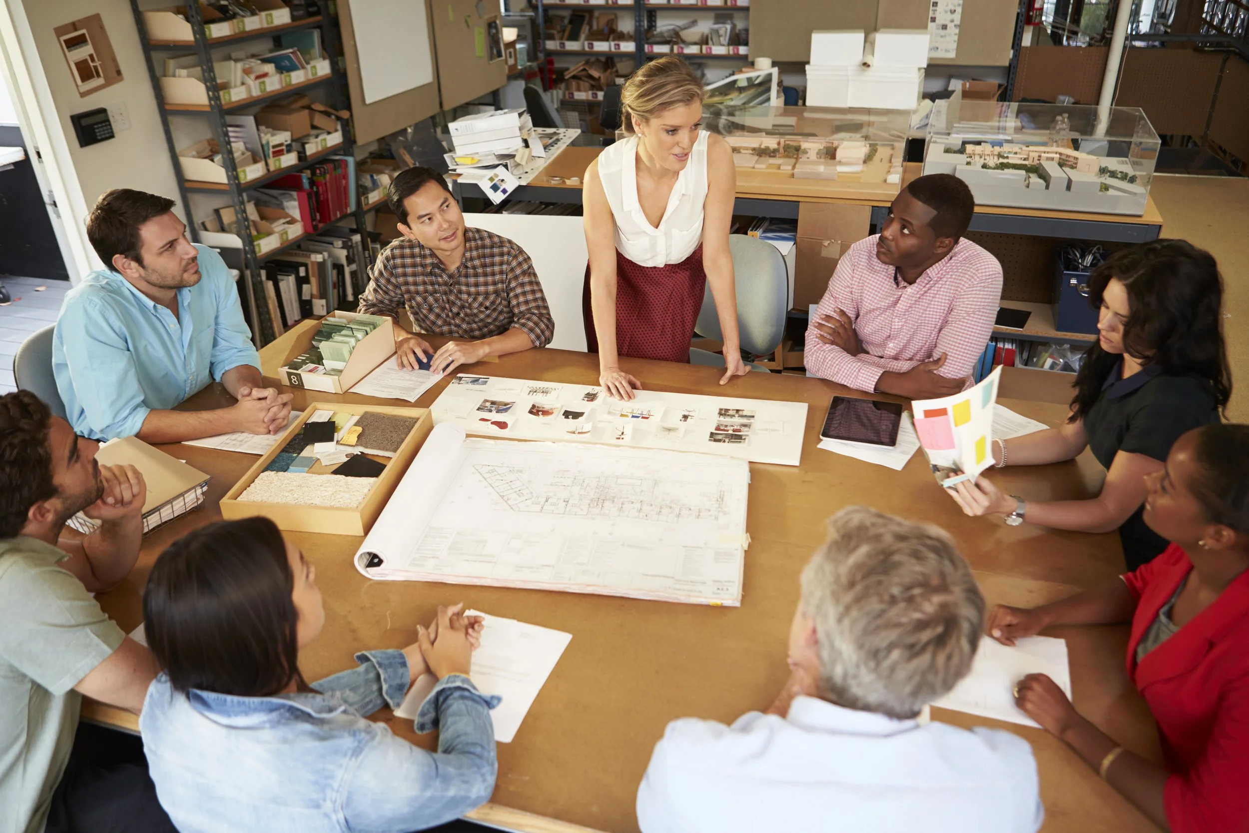 A diverse group of people sit around a table reviewing architectural plans and project materials in a design studio.
