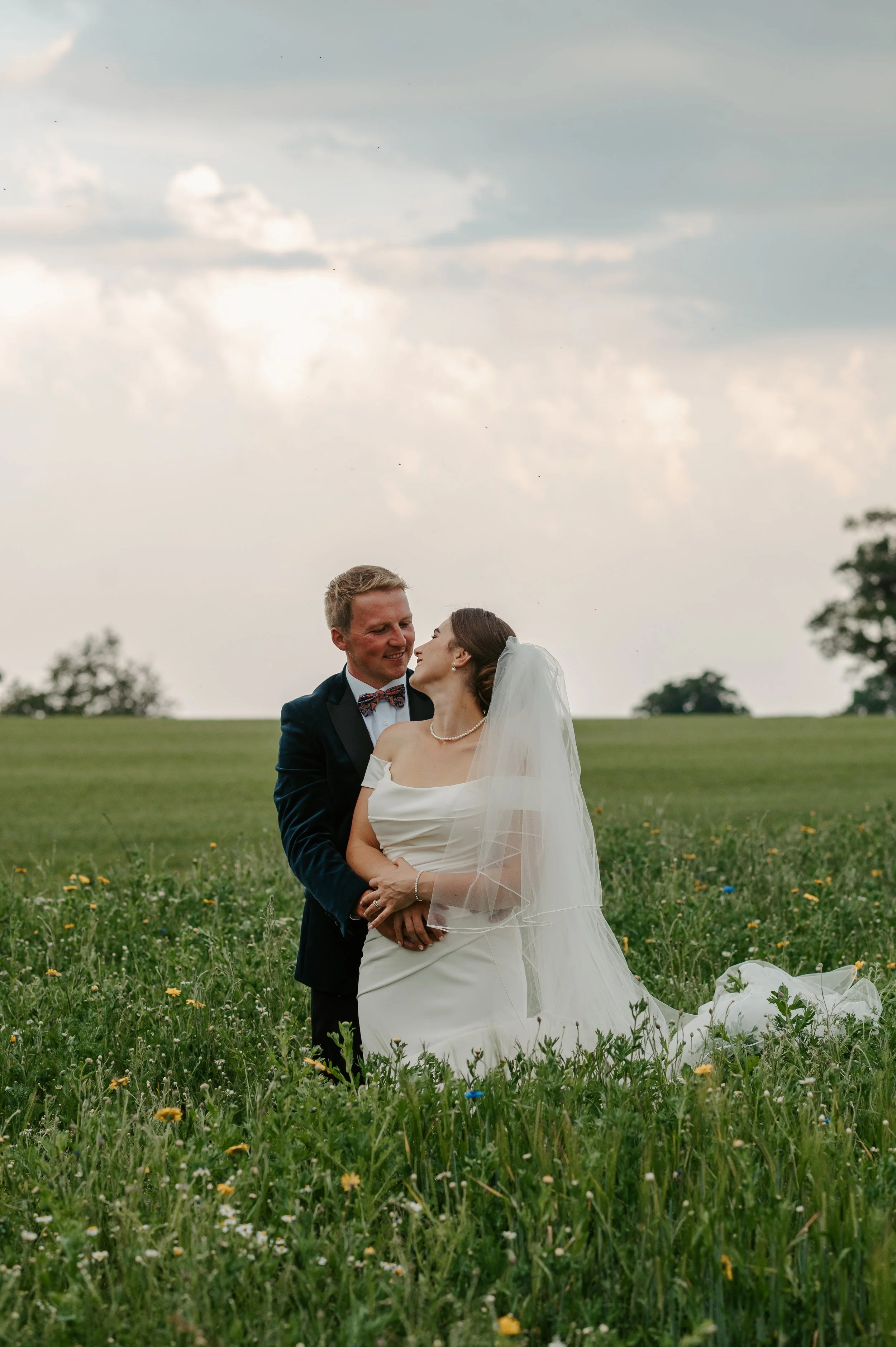 A bride and groom standing together in a grassy field with wildflowers, looking at each other lovingly, under a partly cloudy sky during their wedding. Held at a wedding in Shropshire coordinated by Bootique Weddings and Events.