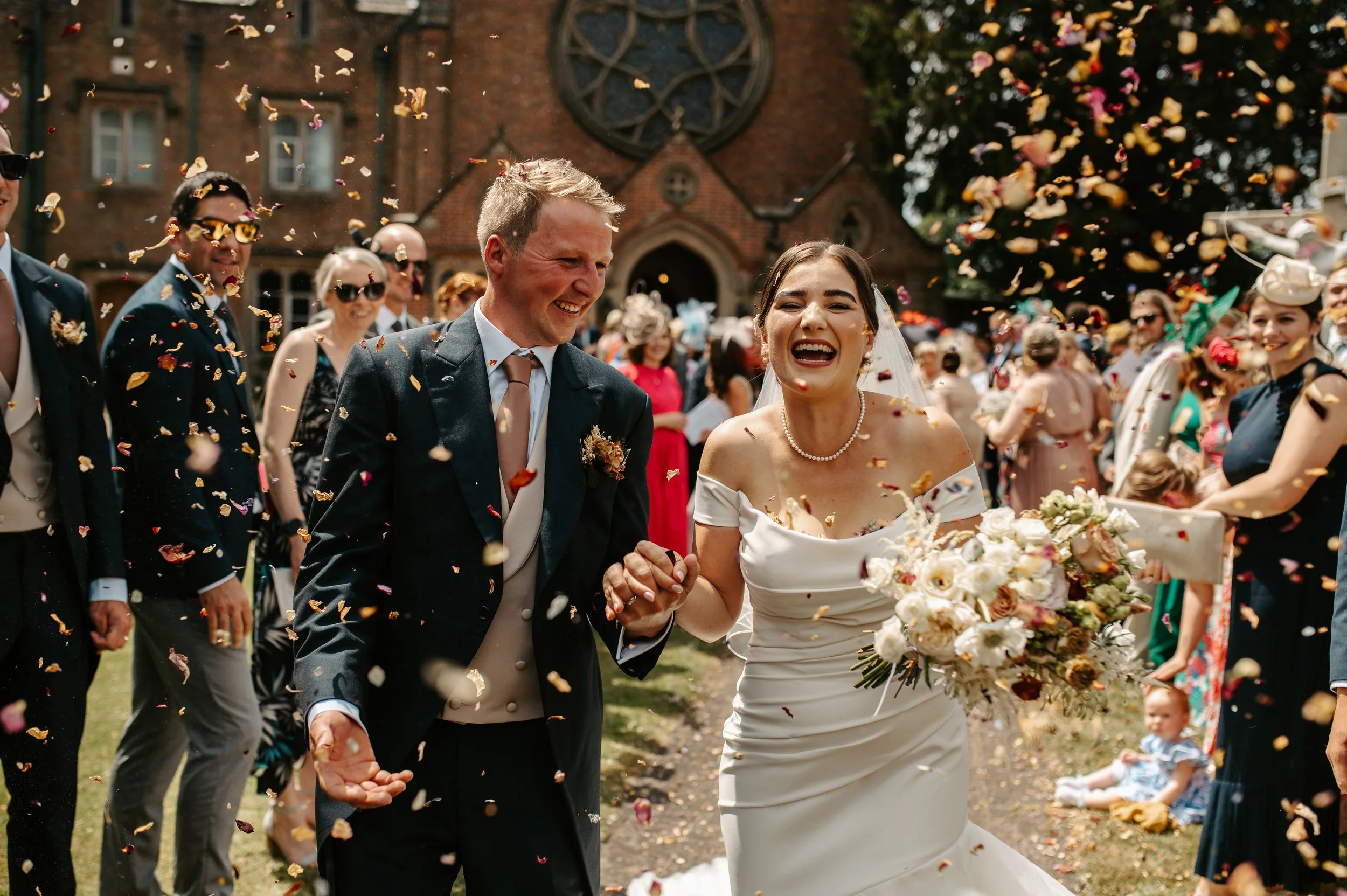 Bride and groom holding hands and smiling as guests throw confetti during wedding celebration outside church. Held at a wedding in Shropshire coordinated by Bootique Weddings and Events.