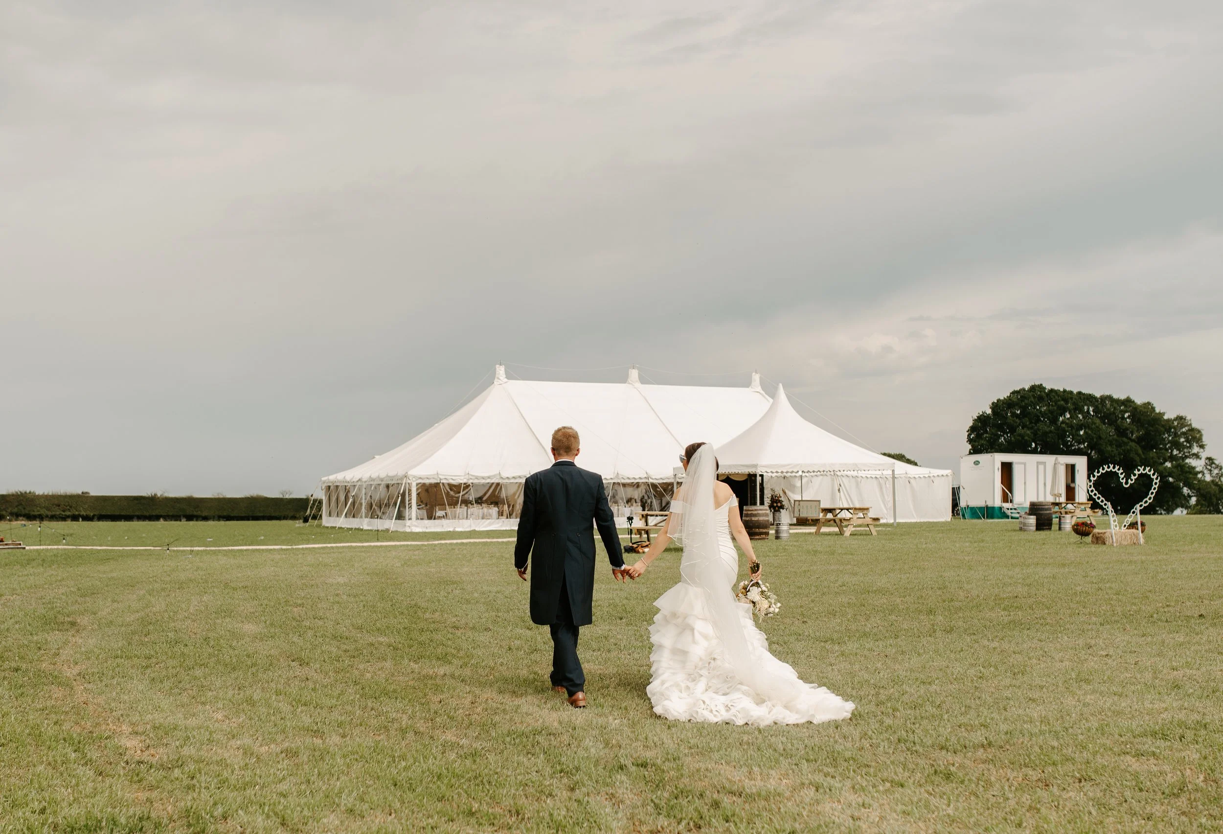 A bride and groom walking hand in hand on a grassy field toward a large white wedding marquee, with wedding decorations and a smaller white trailer in the background. Held at a wedding in Shropshire coordinated by Bootique Weddings and Events.