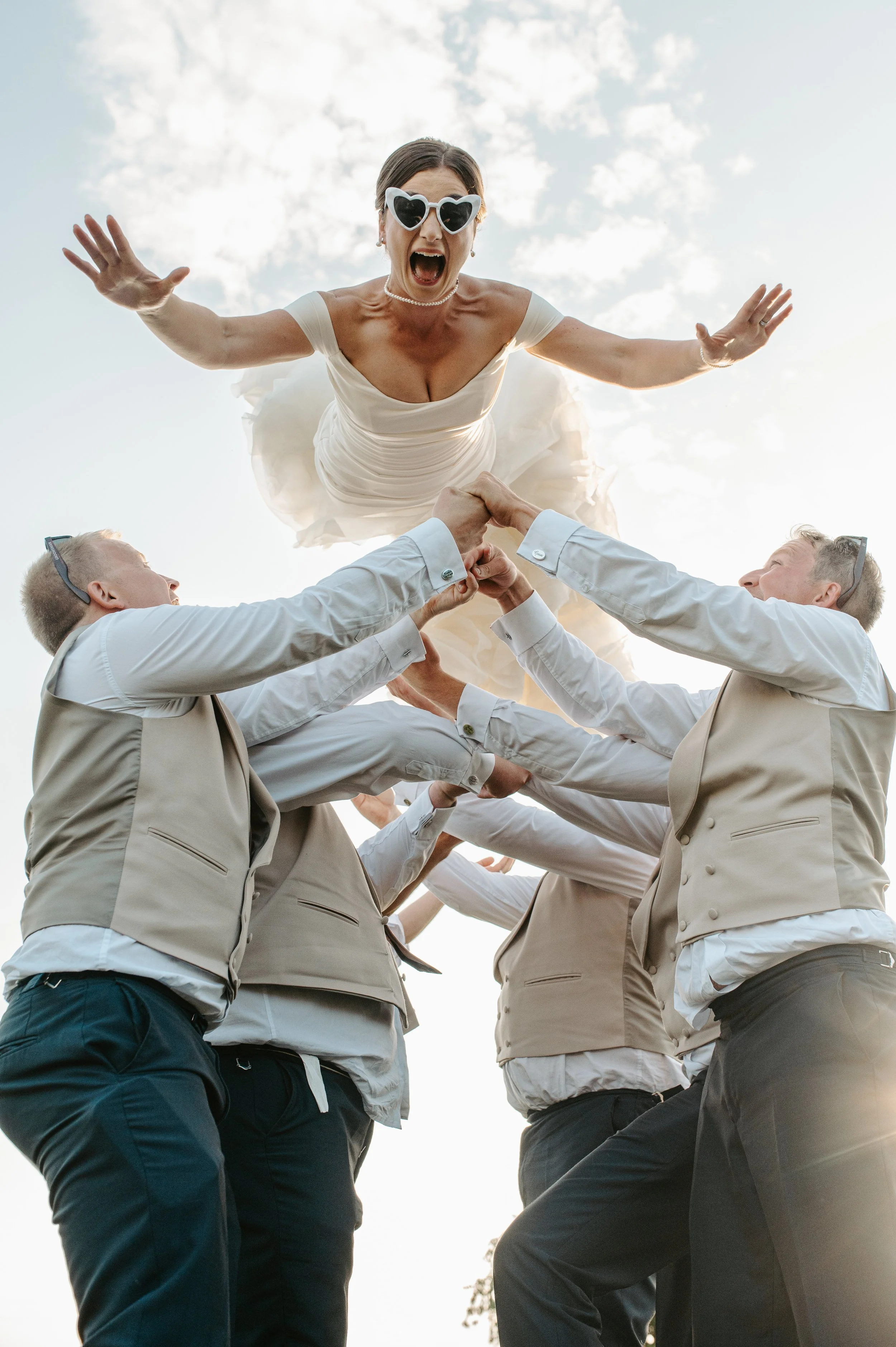 A bride in a white dress with heart-shaped sunglasses is lifted in the air by a group of men in formal attire during an outdoor celebration under a partly cloudy sky. Held at a wedding in Shropshire coordinated by Bootique Weddings and Events.