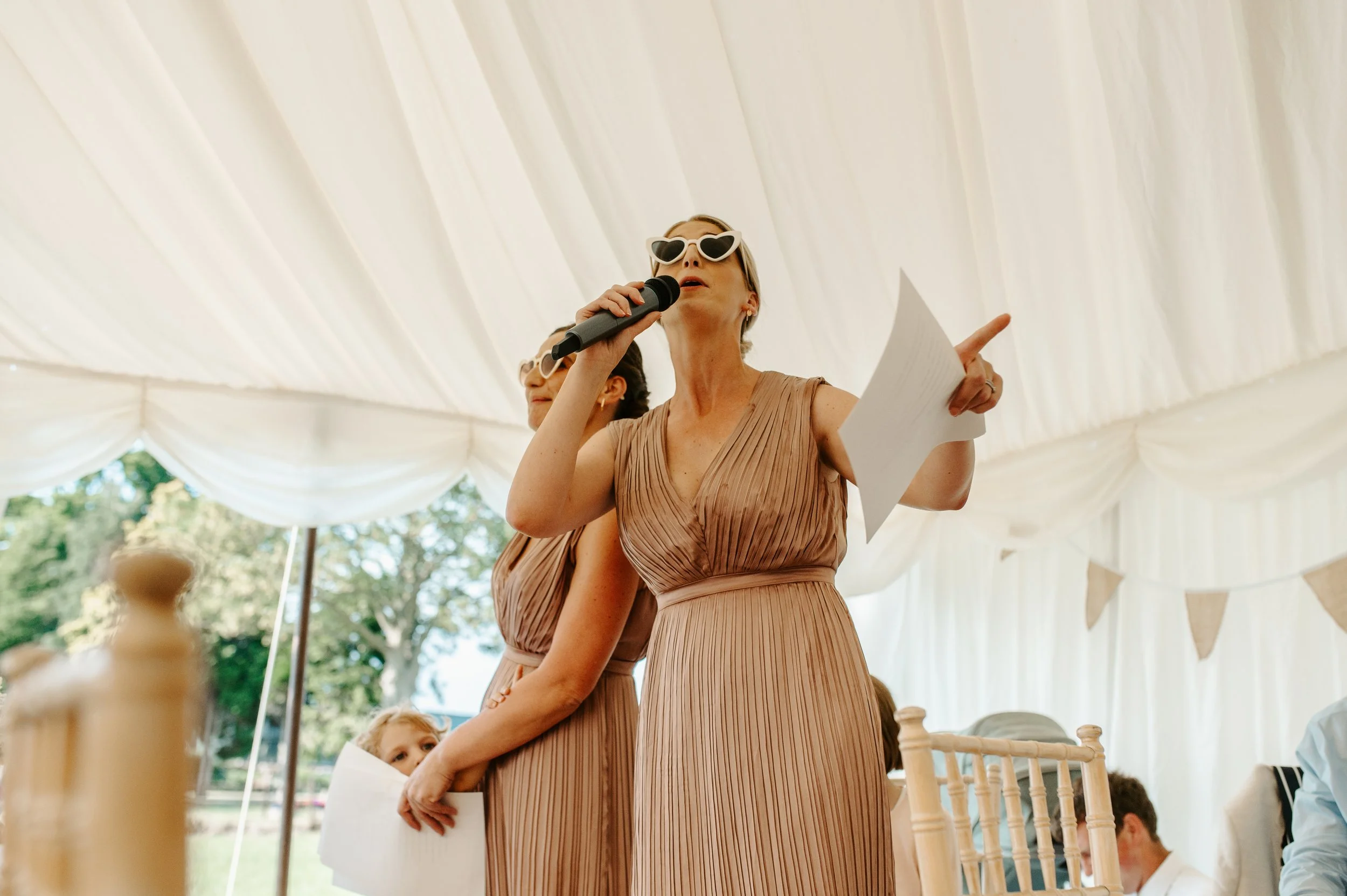 Woman in beige dress with heart-shaped sunglasses speaking into a microphone at a wedding or celebration event, holding papers, under a white marquee with decorative bunting. Held at a wedding in Shropshire coordinated by Bootique Weddings and Events