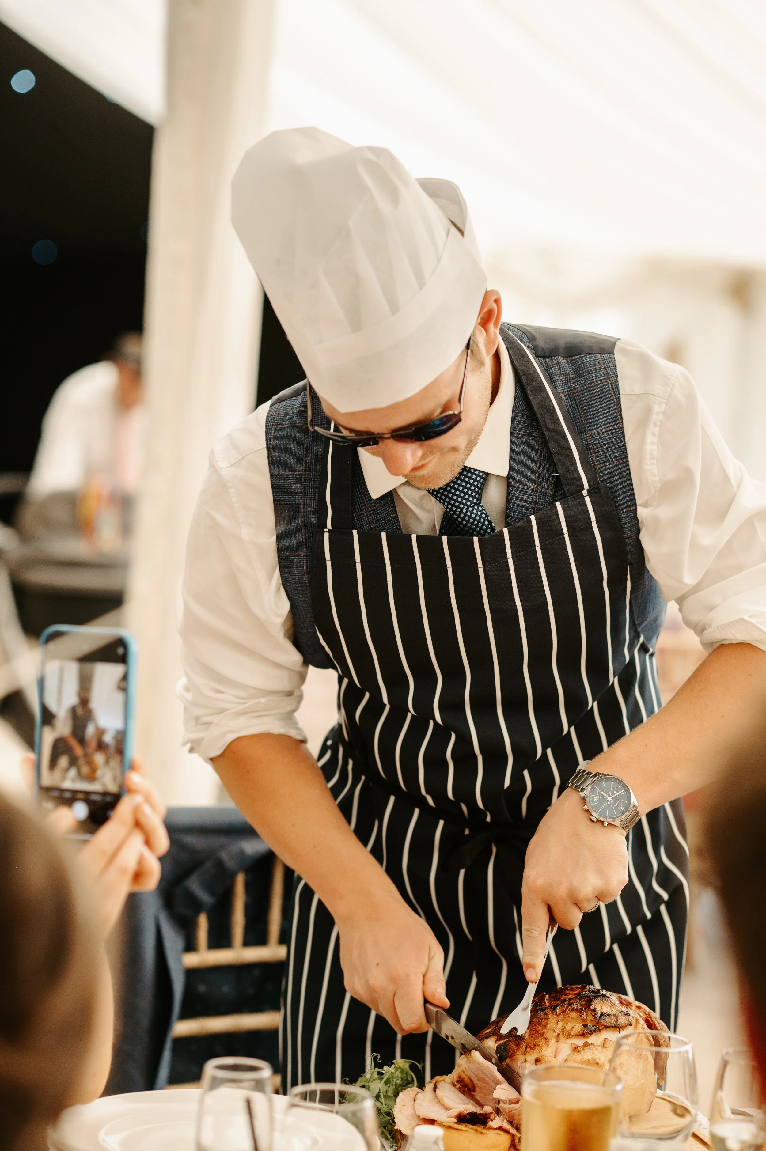 Man dressed as a chef carving a roasted meat at a celebration or event, people taking photos, table with glasses and plates. Held at a wedding in Shropshire coordinated by Bootique Weddings and Events.