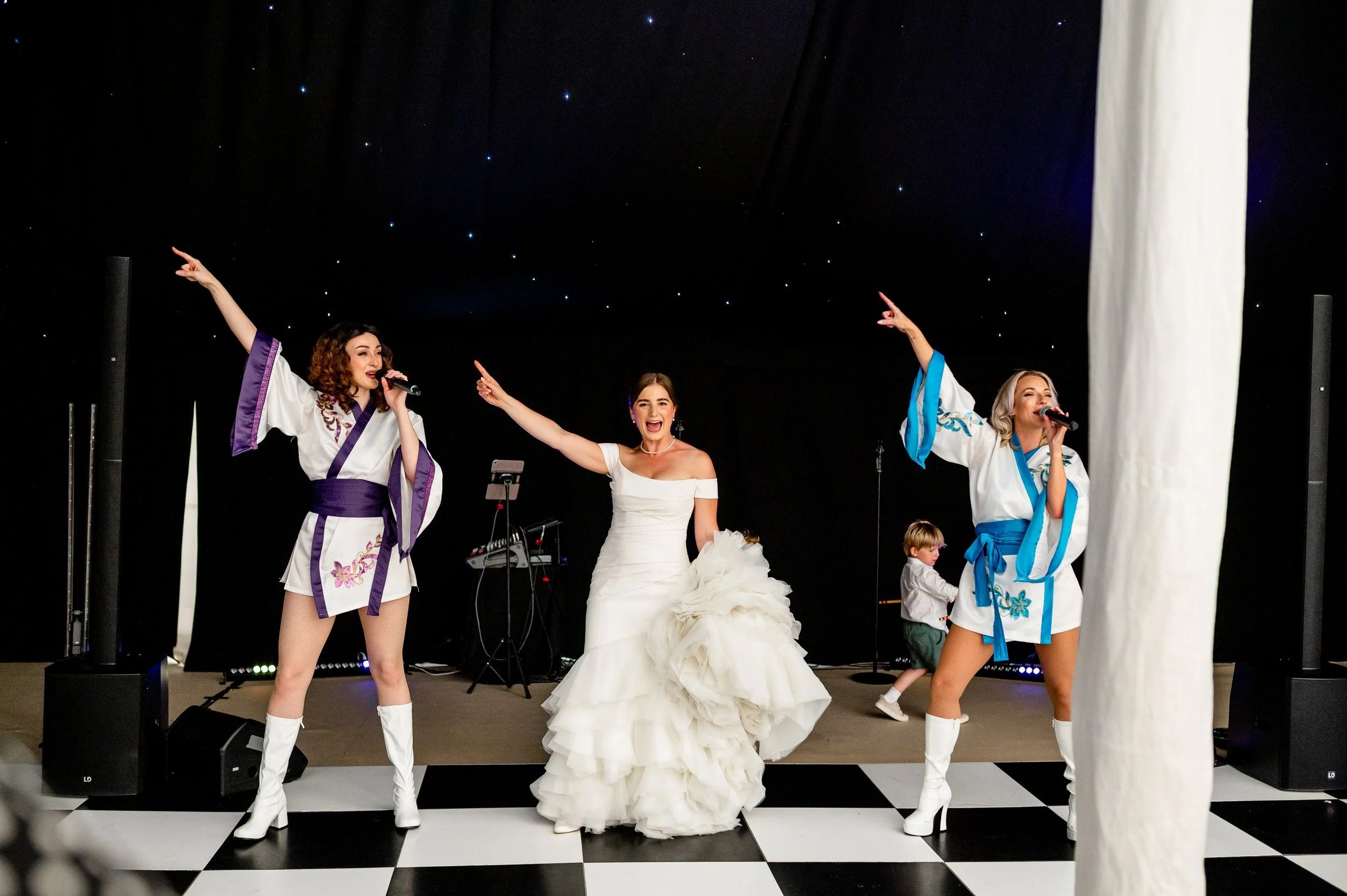 Three women singing and dancing to ABBA songs on a stage with a black background and star-like lights. Held at a wedding in Shropshire coordinated by Bootique Weddings and Events.