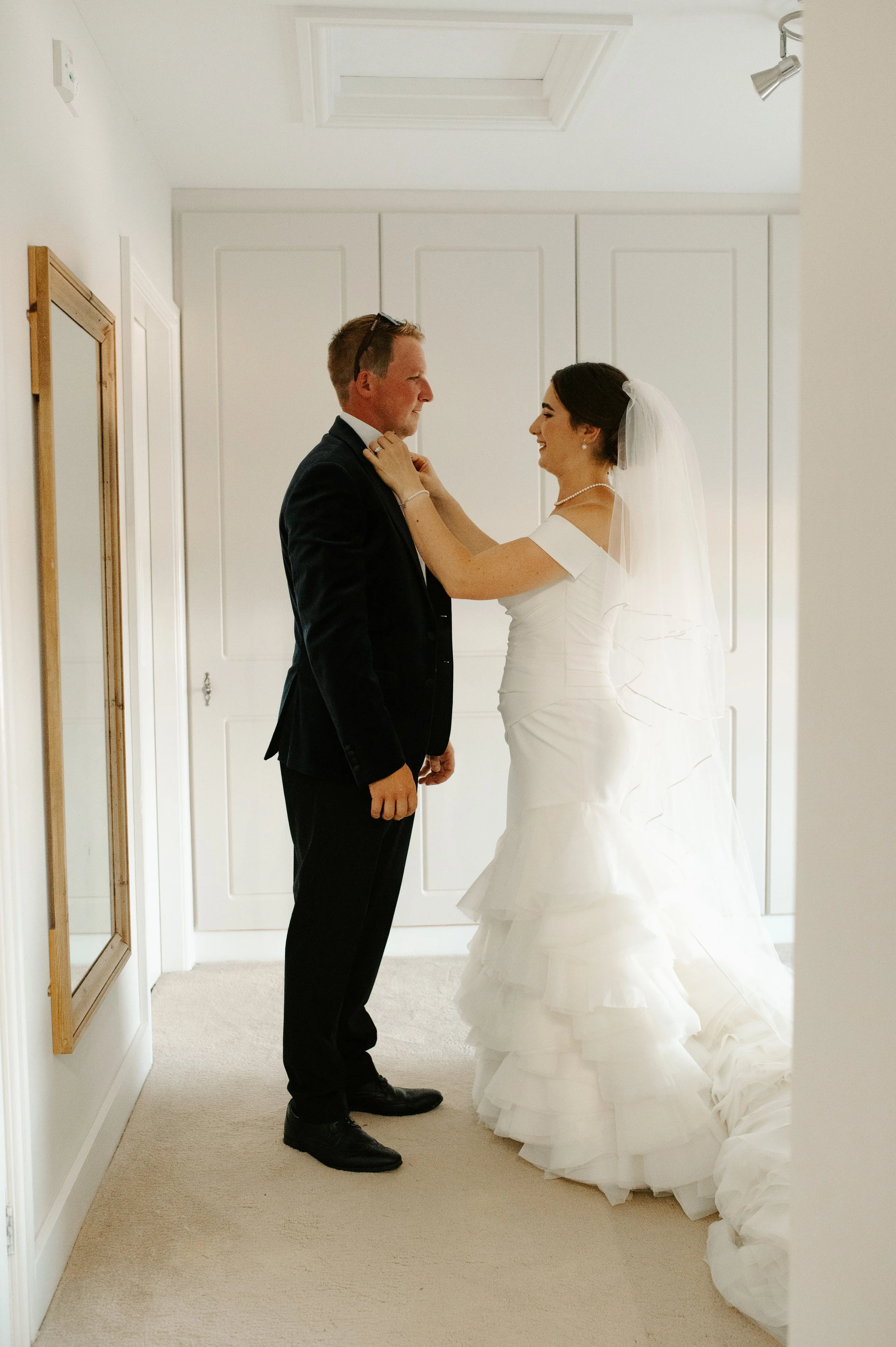 A bride in a white wedding dress and veil is adjusting the groom's tie in a white-room interior. Picture taken at a wedding in Shropshire coordinated by Bootique Weddings and Events.