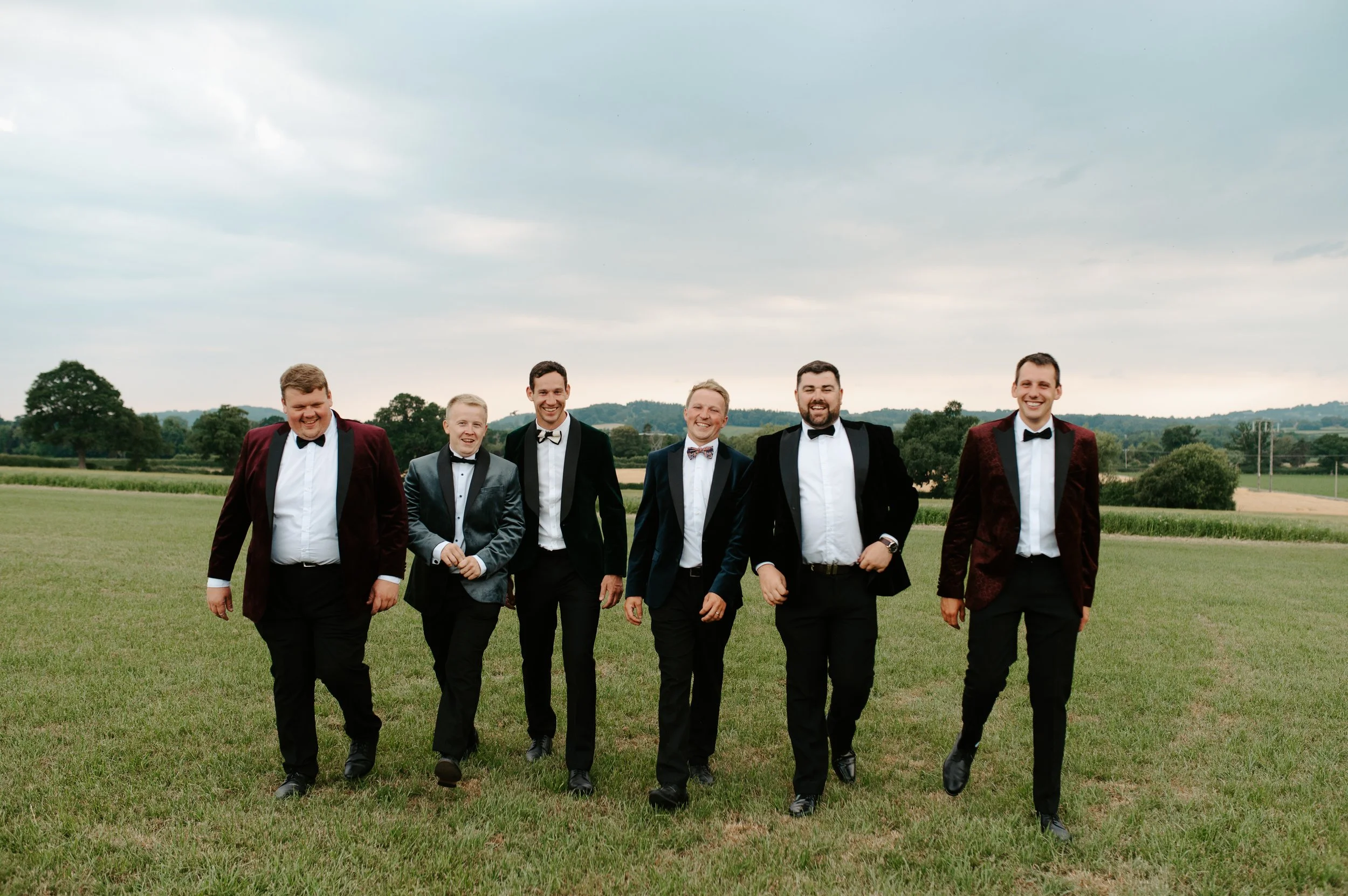 Group of six men in tuxedos walking on a grassy field with trees and cloudy sky in background. Picture taken at a wedding in Shropshire coordinated by Bootique Weddings and Events.