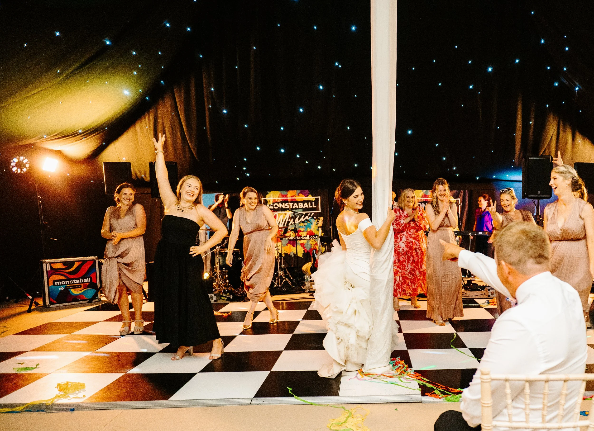 Joyful celebration with women dancing on a checkered dance floor, a woman in a white dress and a man in white shirt pointing, in a decorated black tent with star-like lights and band equipment in the background.
