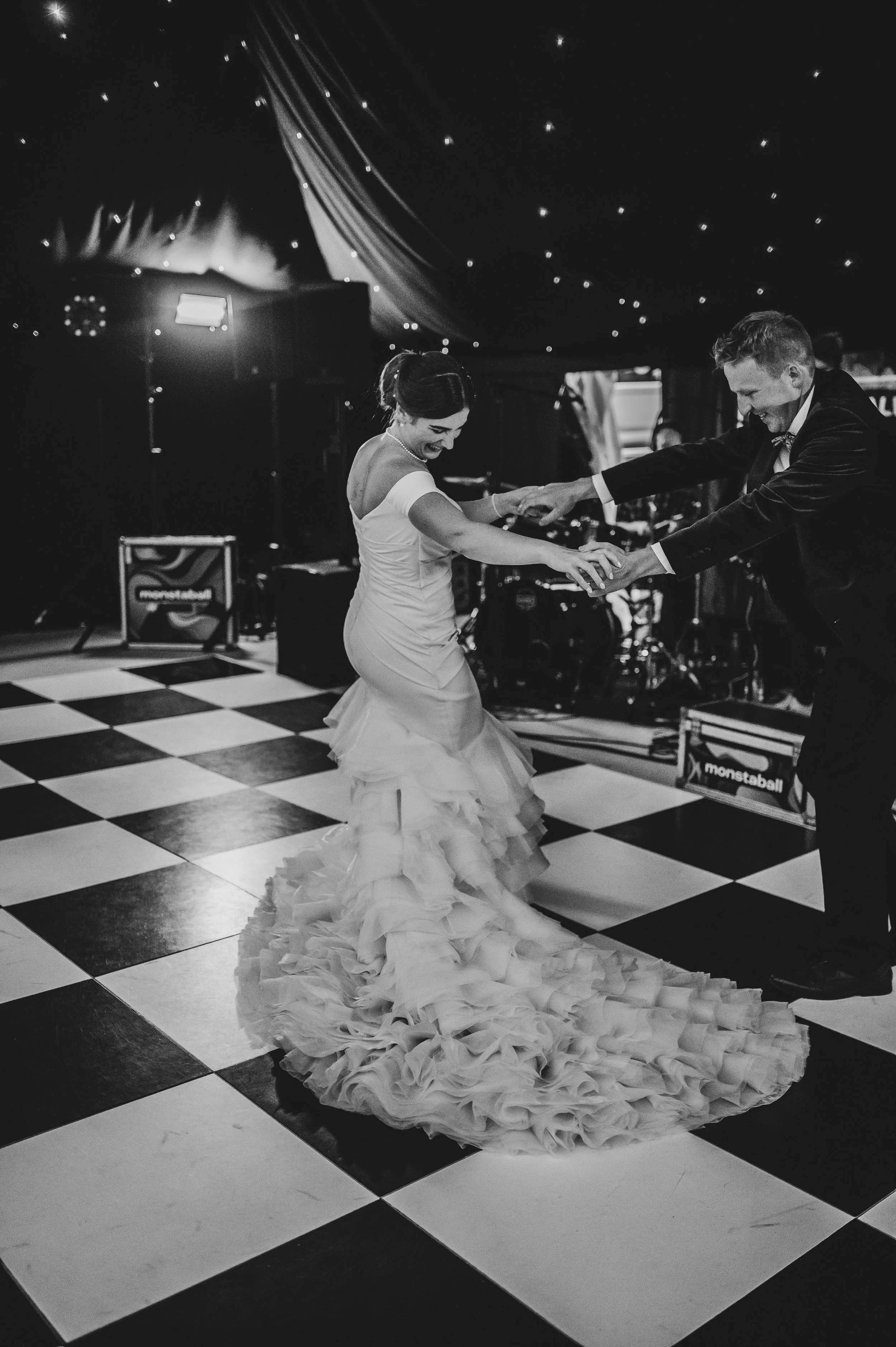 A bride and groom dancing together on a checkered dance floor at their wedding reception, smiling and holding hands, with a band performing in the background. Picture taken at a wedding in Shropshire coordinated by Bootique Weddings and Events.