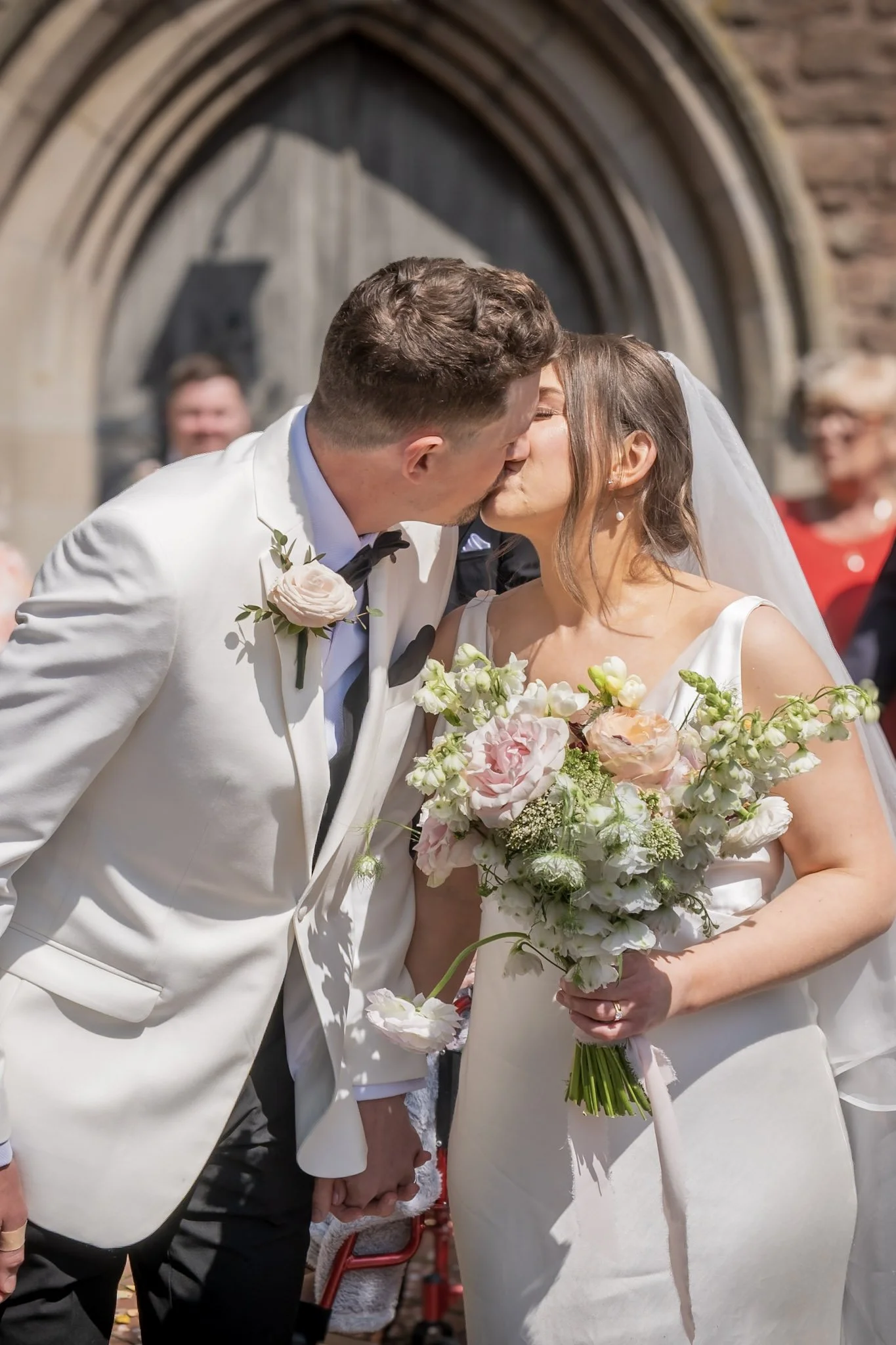 A bride and groom sharing a kiss on their wedding day outside, with the bride holding a bouquet of pink and white flowers, and the groom dressed in a white tuxedo with a black bow tie. Taken outside a church in Shropshire.