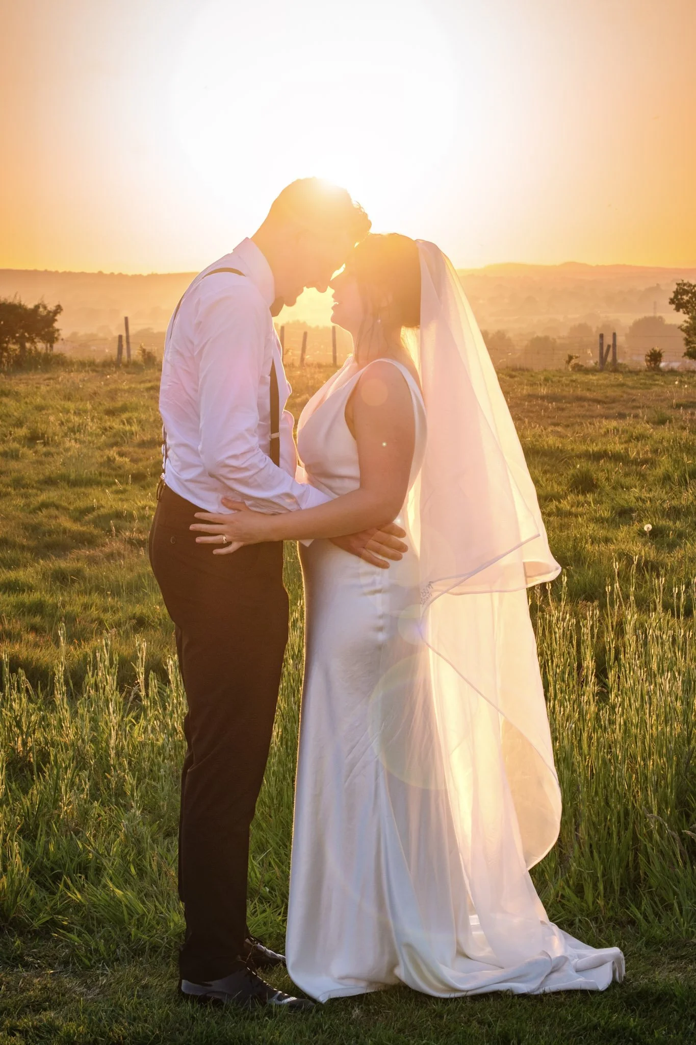 A bride and groom standing close together in a field at sunset, touching foreheads with the sun shining behind them. Held at wedding in Shropshire planned by Bootique Weddings and Events.