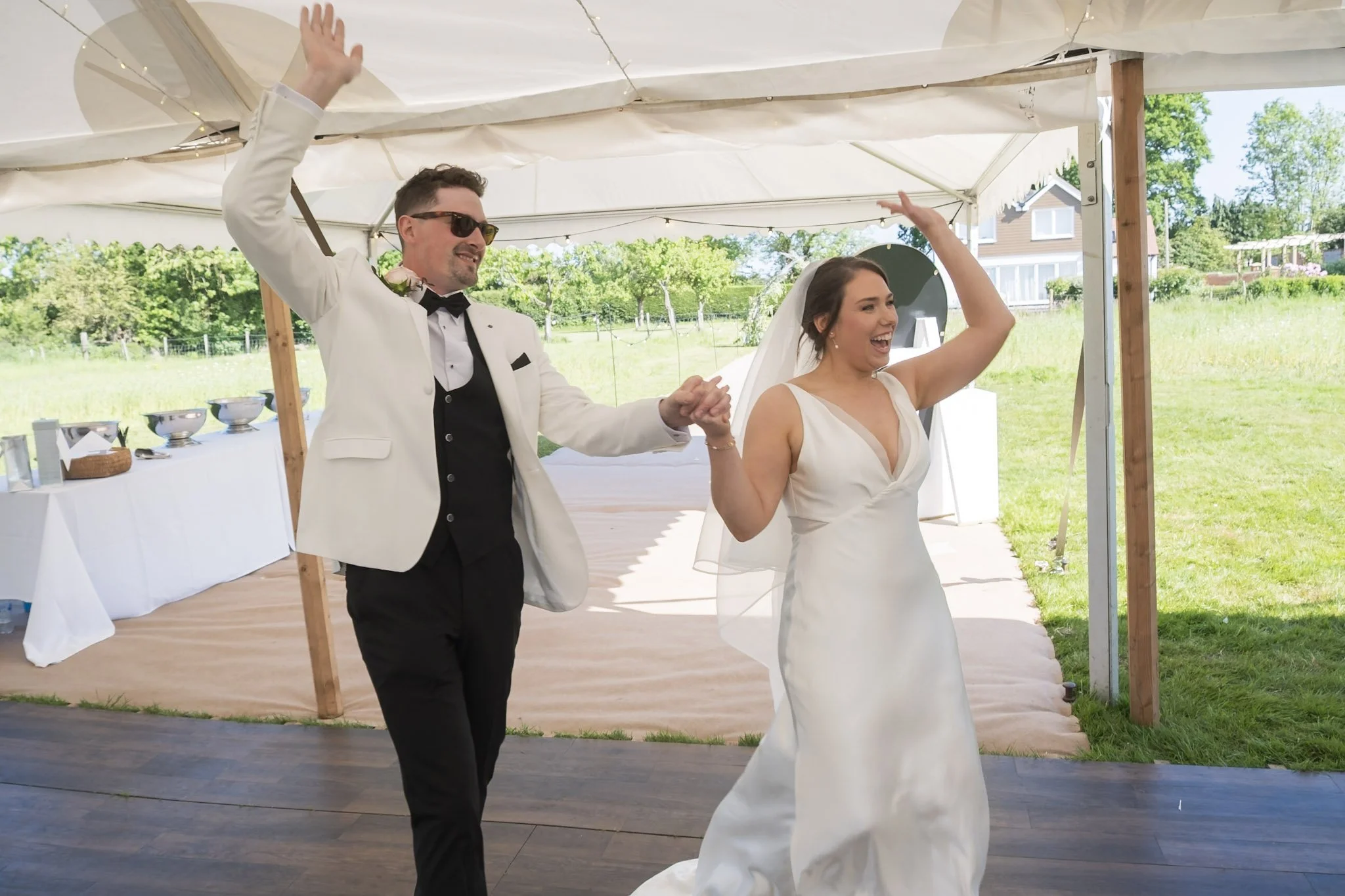 A bride and groom entering happily with their hands in the air under a white marquee at their wedding reception in Shropshire, planned by Bootique Weddings and Events.
