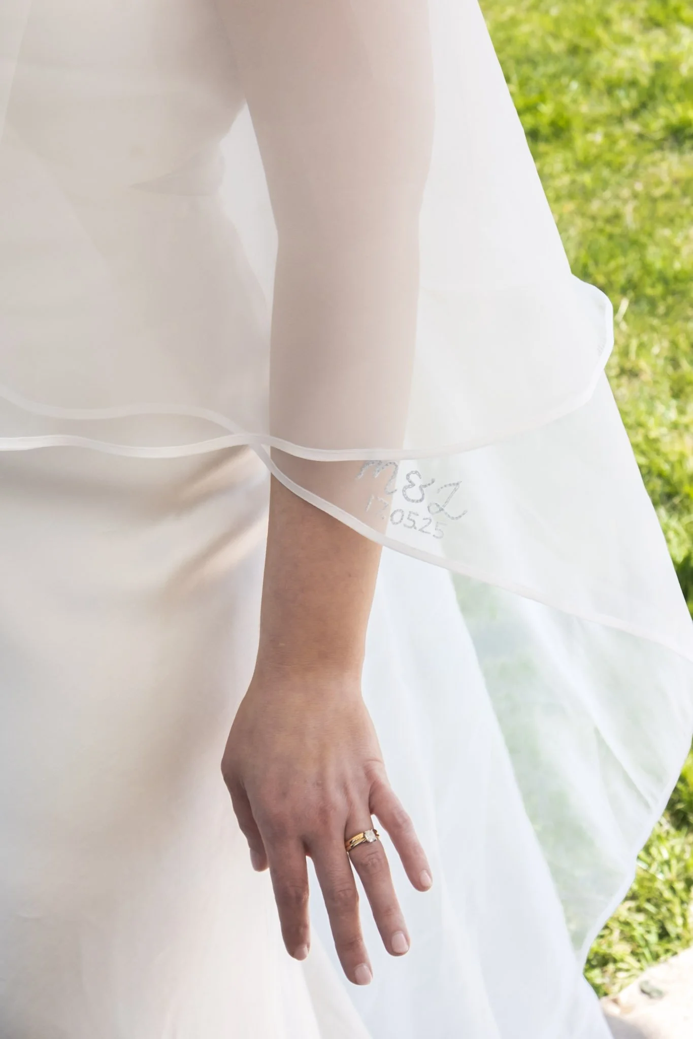 Close-up of a bride's hand with wedding rings, wearing a wedding dress and sheer veil, outdoors on grass.