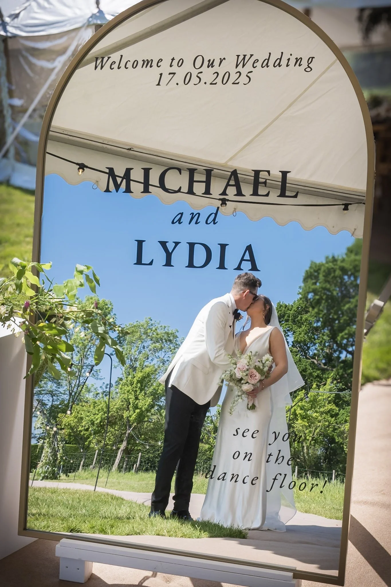 A wedding sign with a mirror reflects a bride and groom kissing outdoors, under a tent, with trees and a blue sky in the background. Held at a wedding in Shropshire coordinated by Bootique Weddings and Events.