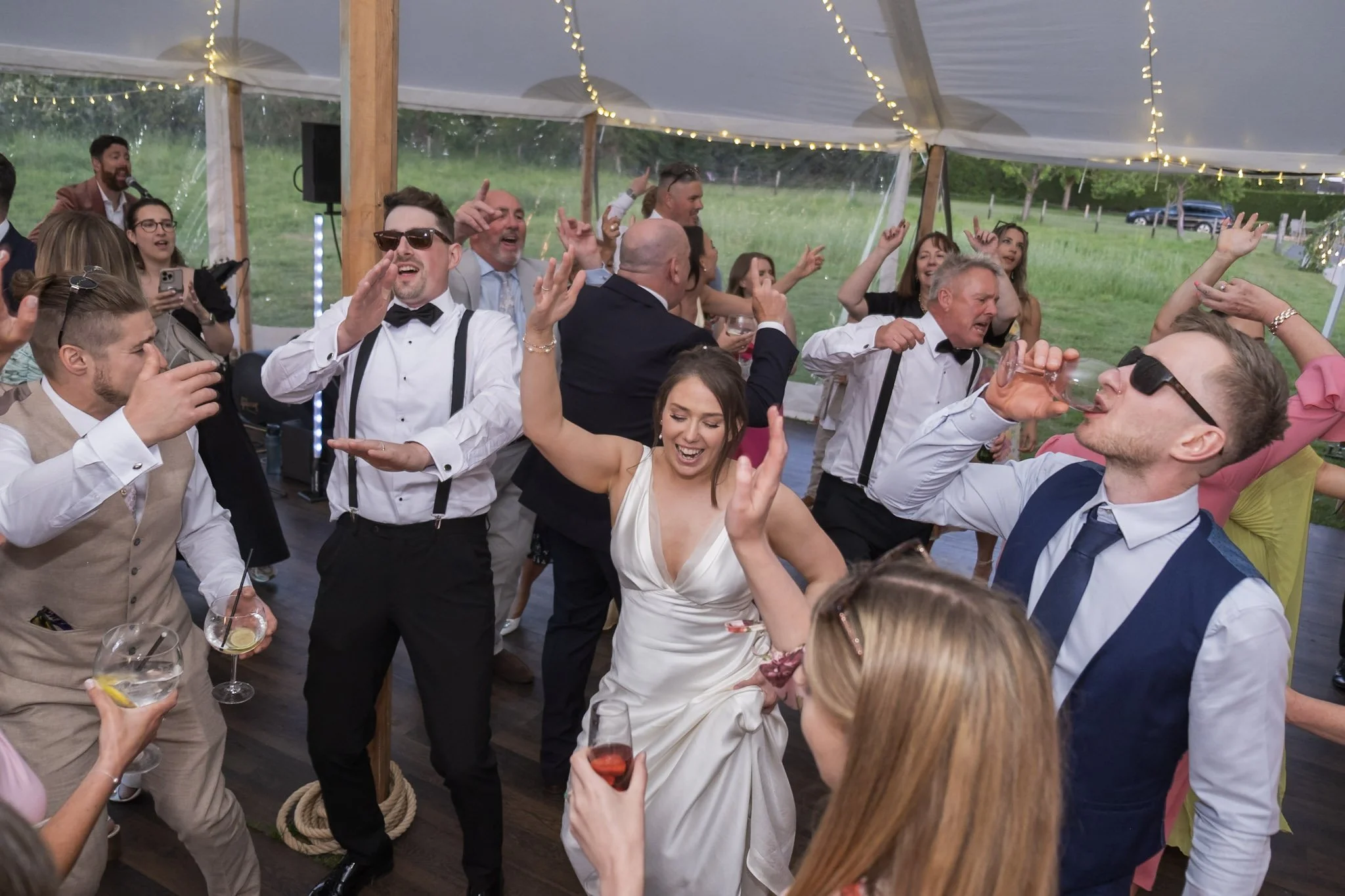 People dancing and celebrating at a wedding reception under a tent, with string lights overhead and a grassy field in the background. Held at a wedding in Shropshire coordinated by Bootique Weddings and Events.