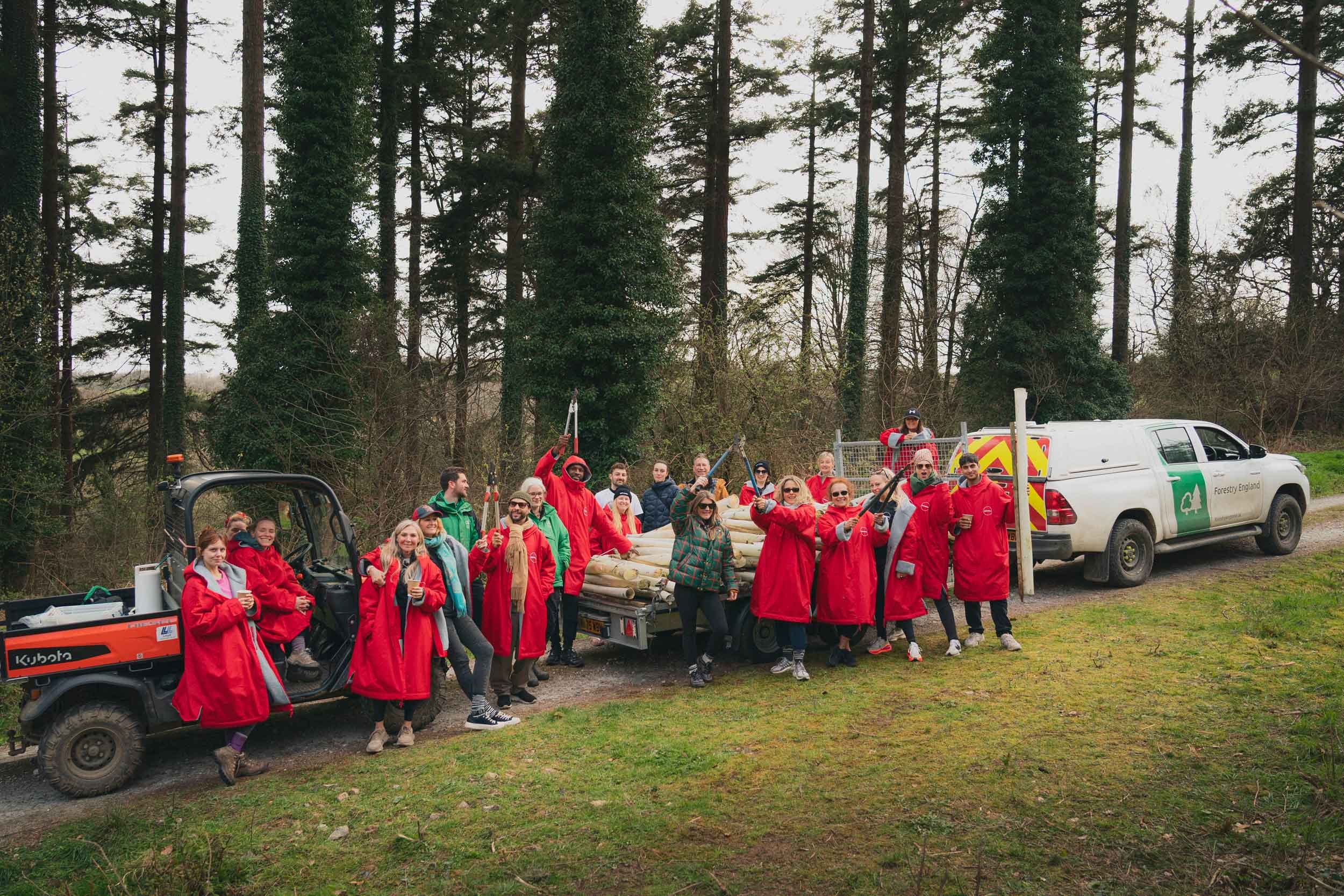 A group of people in outdoor gear gathered around a flatbed truck with logs, in a forested area, participating in a logging or forestry event. Some are holding tools, and the group includes men and women of diverse ages.