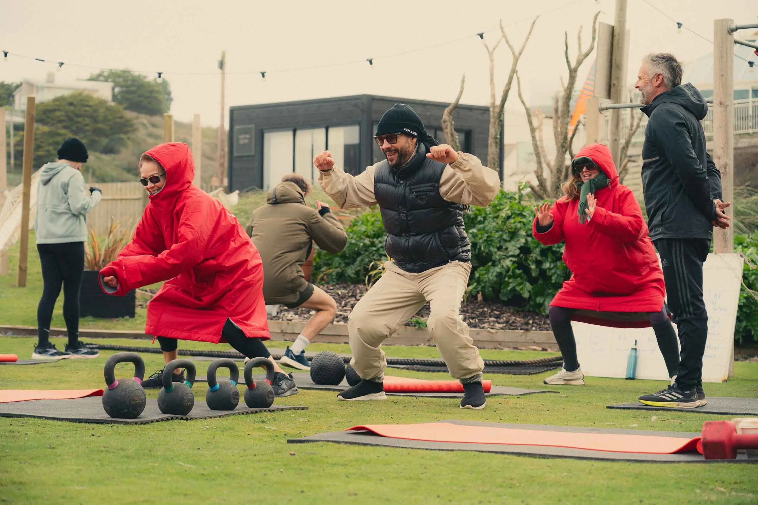 Group of people participating in outdoor fitness class with kettlebells, mats, and orange exercise bands, in a grassy area with trees and a small black building in the background.