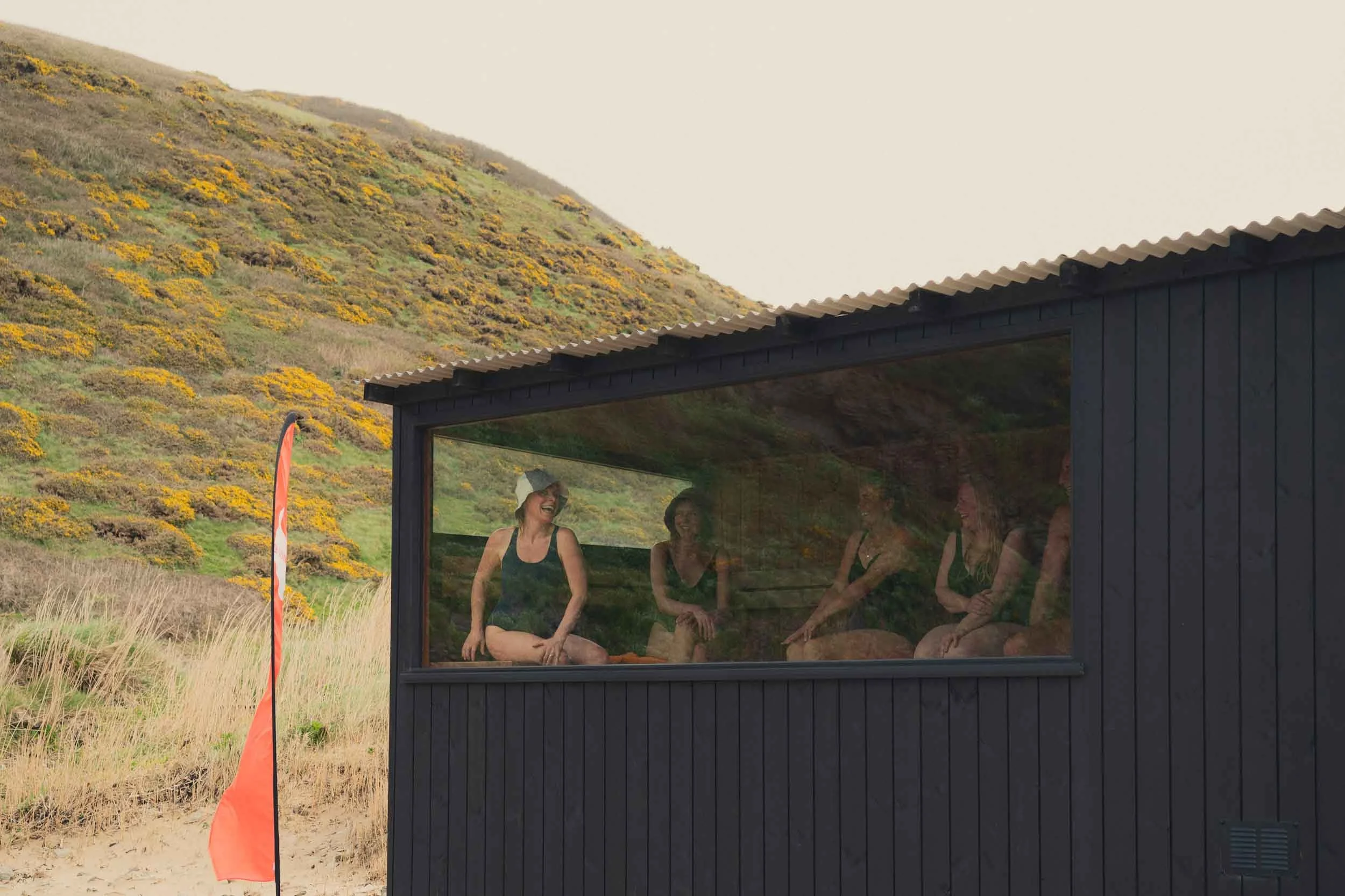 Four women sitting inside a black wooden shed with large glass windows, laughing and engaging with each other, with a backdrop of yellow flower-covered hillside and an overcast sky.