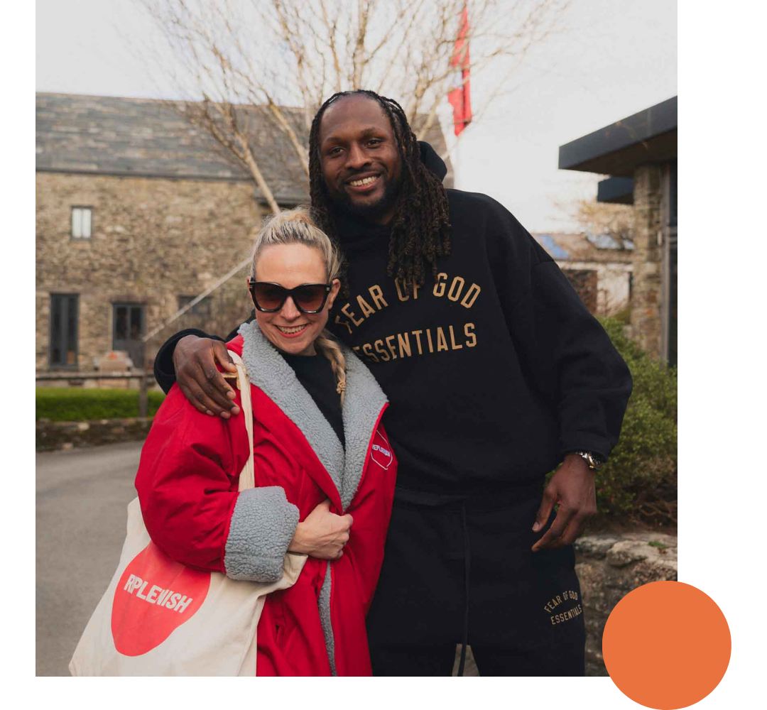 Two people smiling and hugging outdoors. The woman is wearing sunglasses and a red jacket with grey fleece lining, carrying a tote bag that says 'REPLENISH'. The man is wearing a black hoodie with the phrase 'FEAR OF GOD ESSENTIALS' printed on it.