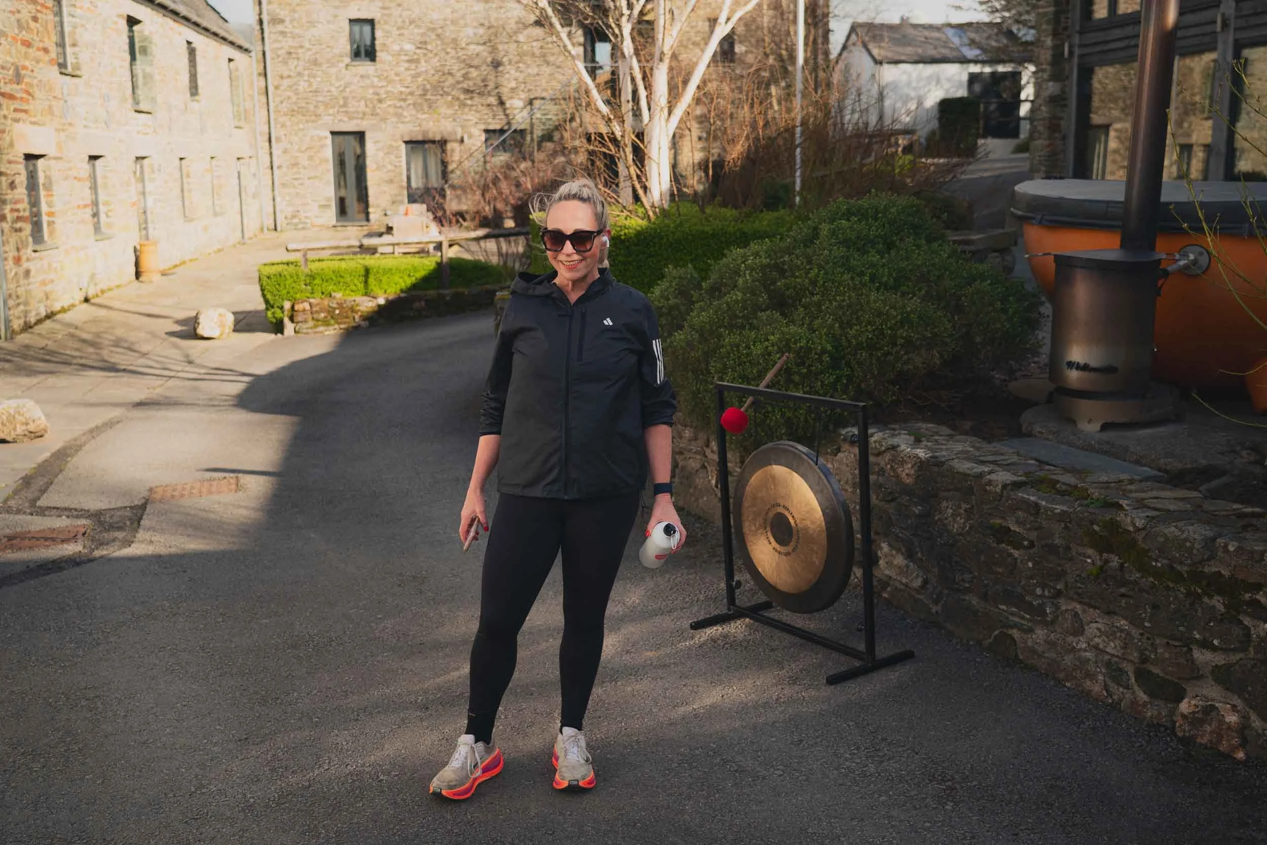 Woman in black athletic wear and sunglasses smiling outdoors, holding a white portable speaker, standing next to a gong and a temple bell, with stone buildings, trees, and bushes in the background.