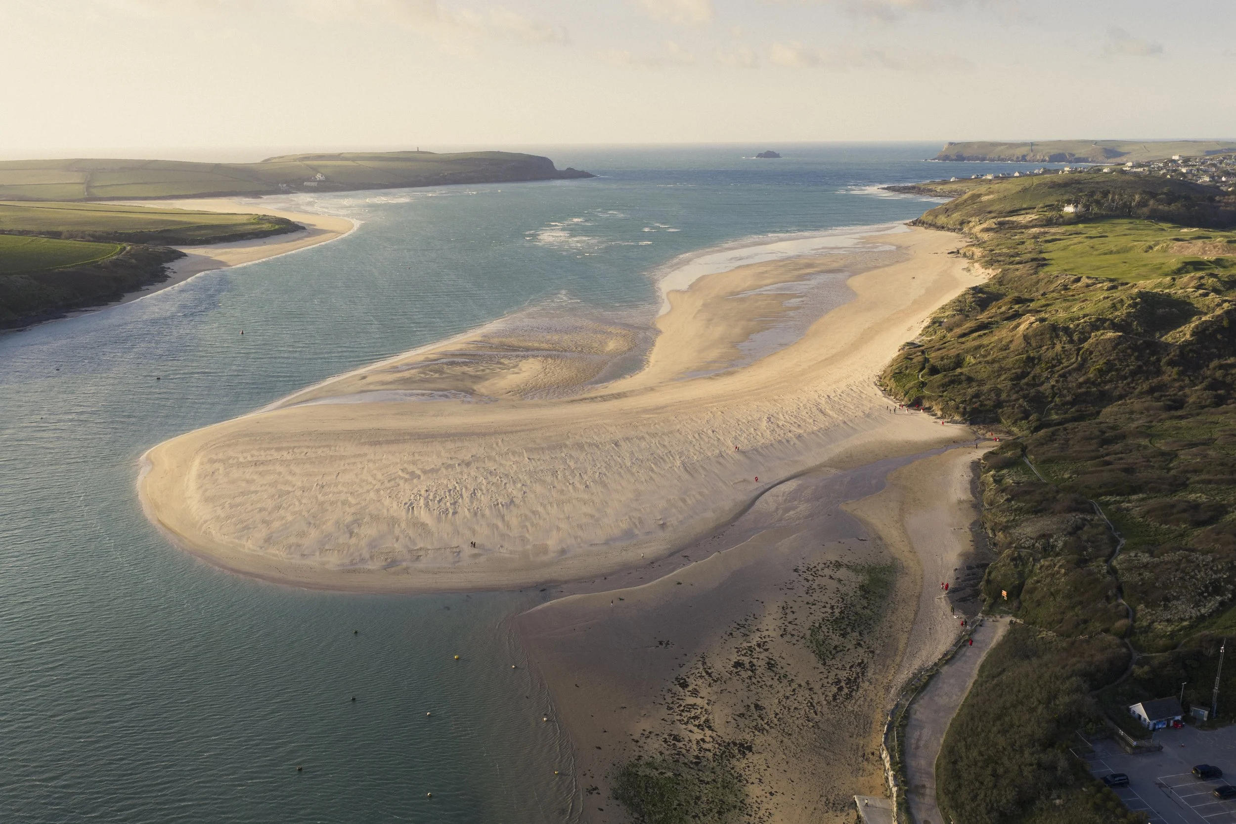 A scenic aerial view of a coastal beach with golden sands, water channels, and green hills on both sides, with a small town visible in the distance on the right.