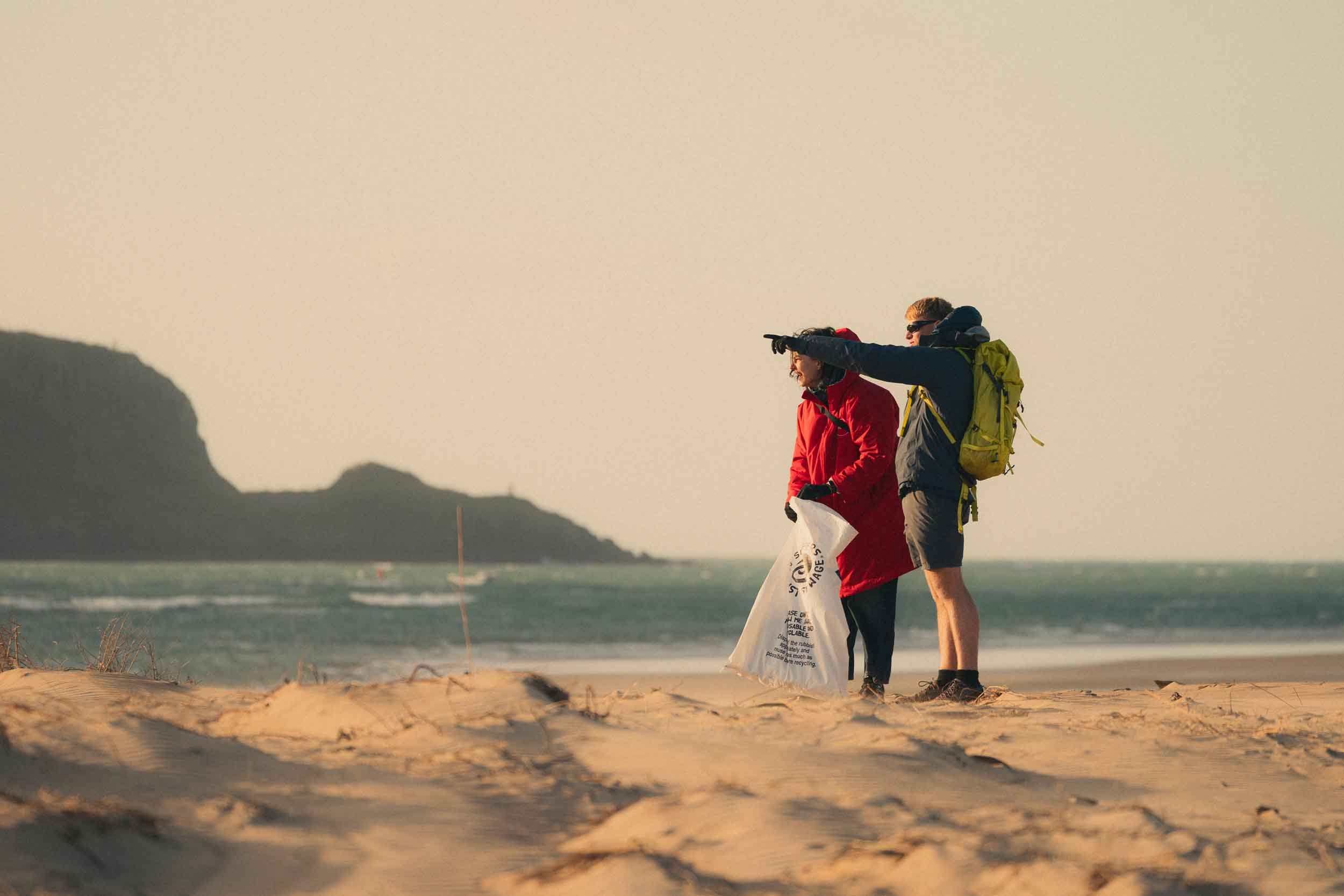 Two young men stand on a beach near the water, with one pointing to the distance. They are dressed in casual outdoor clothing, and one has a yellow backpack. The scene is set during sunset or sunrise, with soft warm lighting and hills or cliffs in the background.