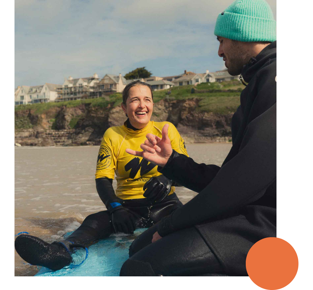 A young woman in a yellow sports jersey and black wetsuit sitting on the beach, smiling at a man in a black jacket and turquoise beanie who is talking to her. They are near the water with houses and cliffs in the background.