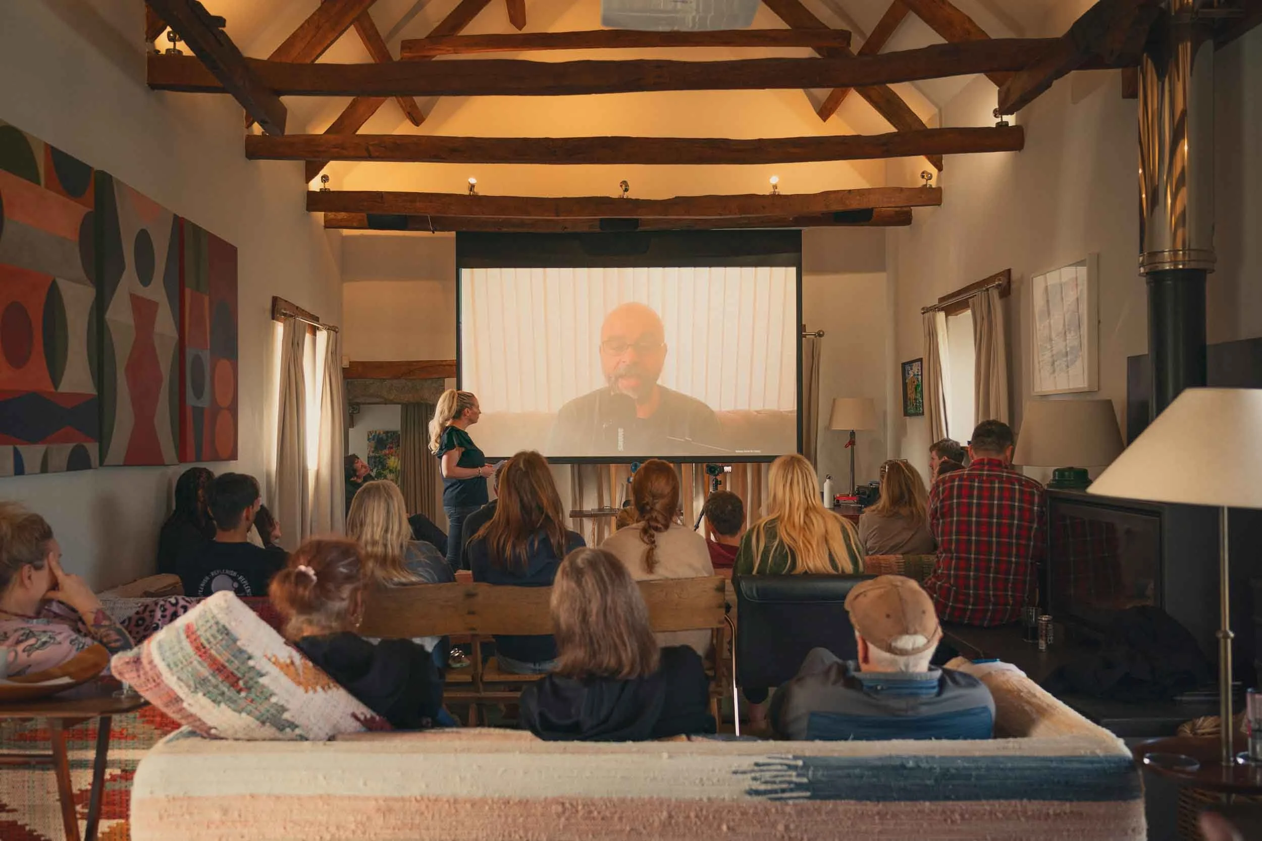 A group of people attending a presentation or lecture in a cozy room with wooden beams, artwork on the walls, and a large screen displaying a man speaking.