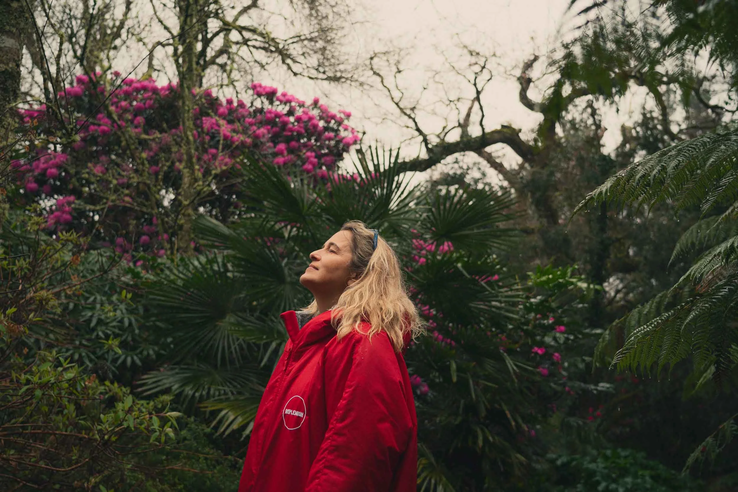 A woman with blonde hair, wearing a red jacket, standing in a lush garden with pink flowers and green foliage, smiling with eyes closed.