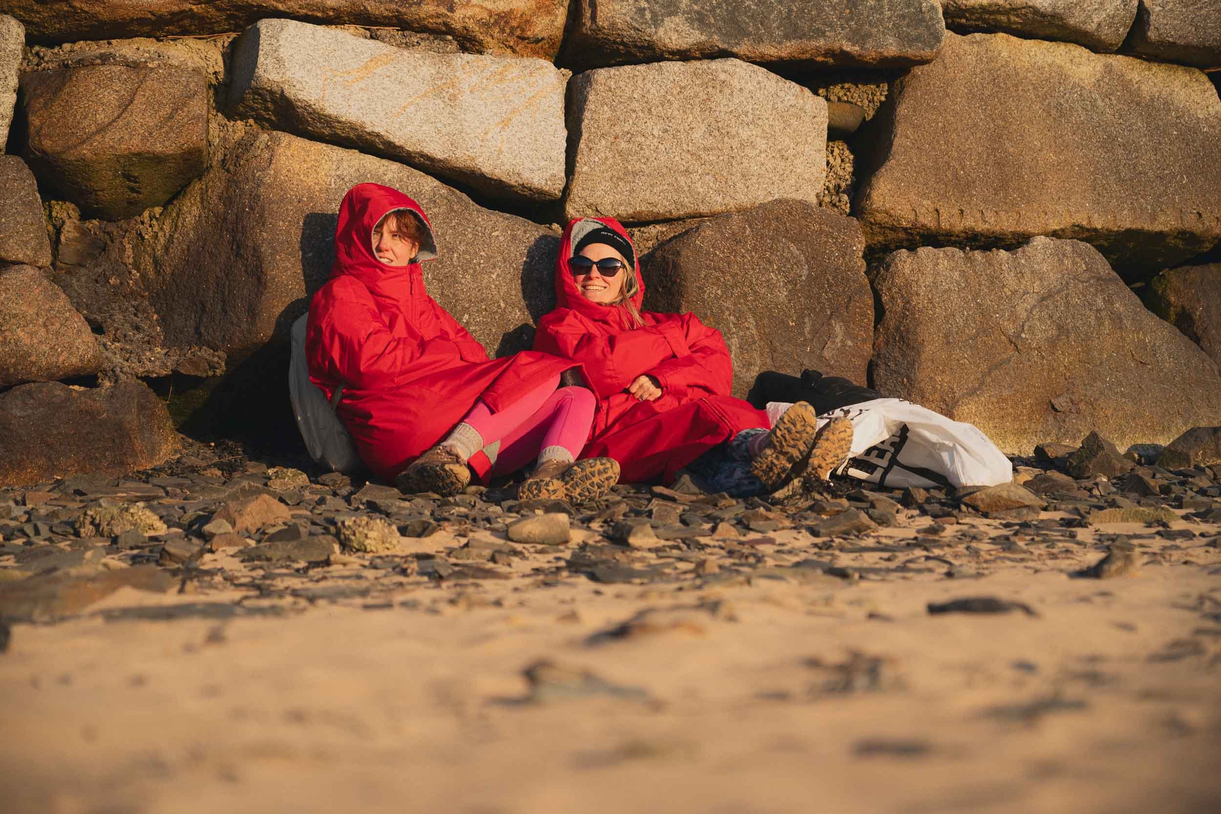 Two women in red jackets and sunglasses sitting on rocks and relaxing on a rocky beach during sunset.