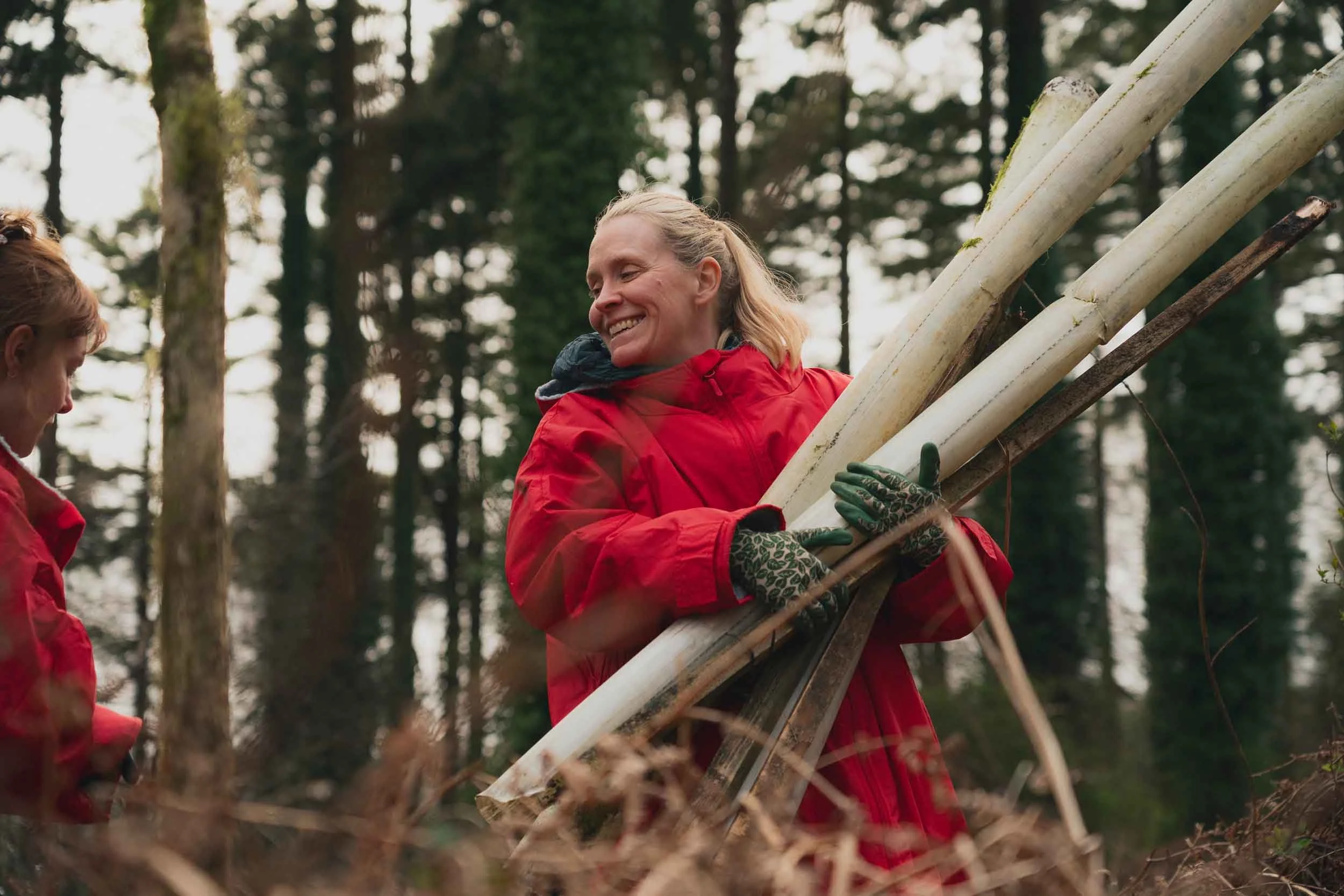 Two women in red jackets working together outdoors in a forest, carrying cut logs on their shoulders and smiling.