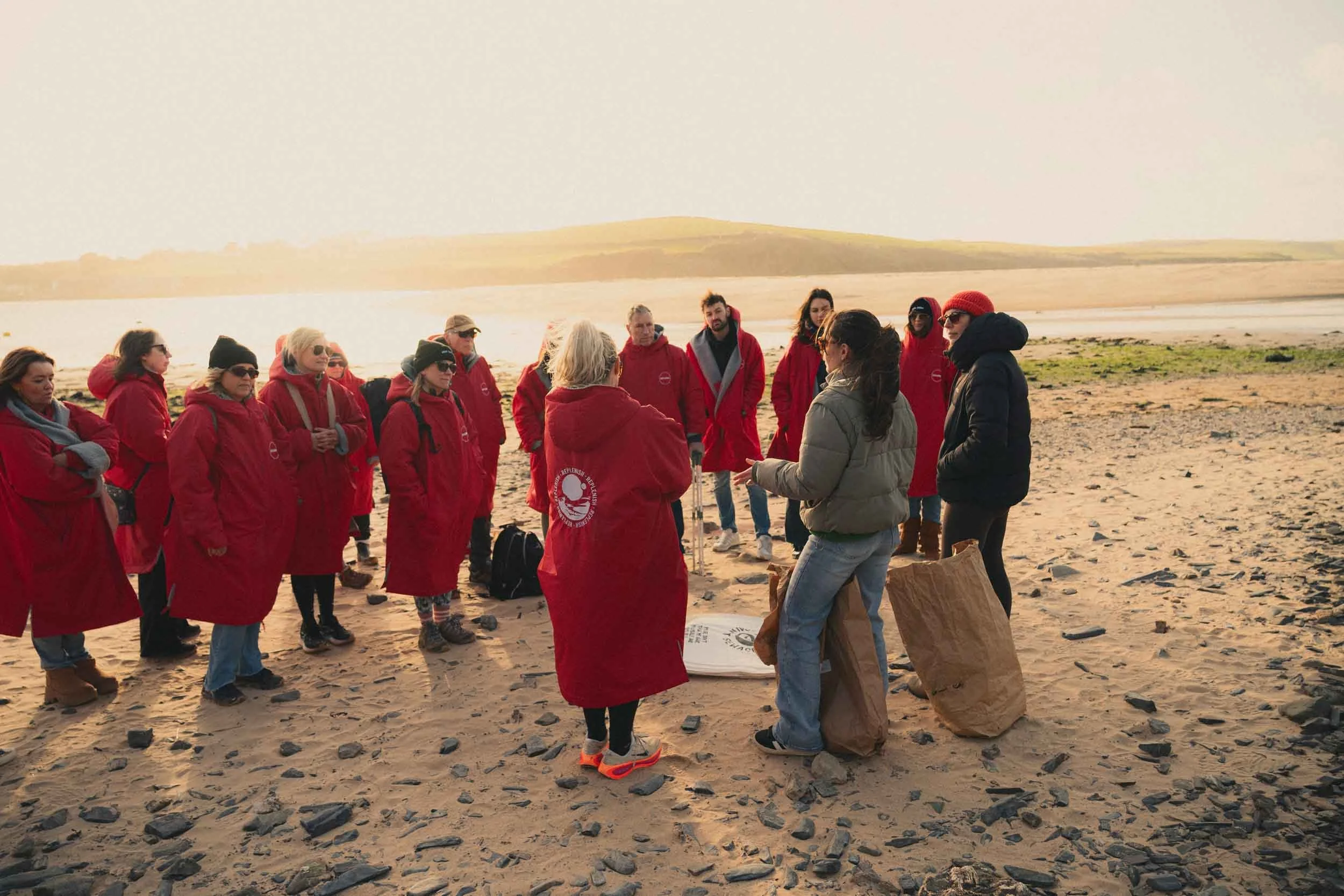 Group of people wearing red jackets listening to a woman speaking on a beach during sunset.