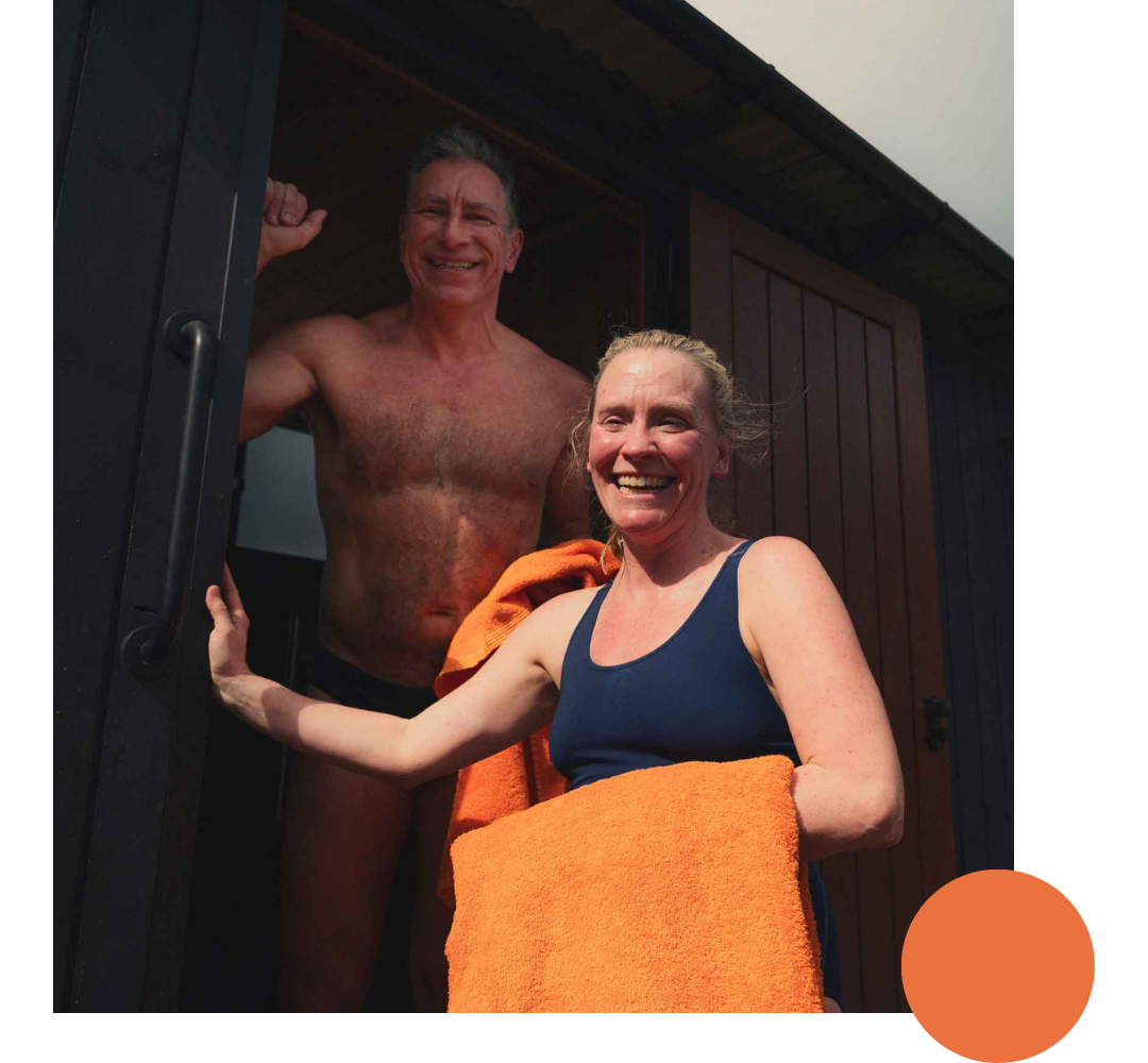 A man and woman stand in a sauna, smiling. The man is shirtless and raising his fist in a celebratory pose. The woman is wearing a navy blue top, holding an orange towel, and standing outside the sauna.