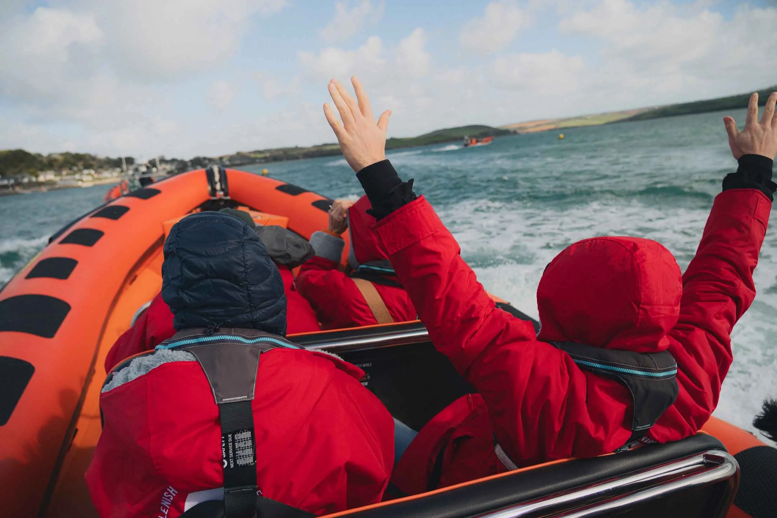 People wearing red jackets riding in an orange inflatable boat on water, waving their hands, with a shoreline and cloudy sky in the background.